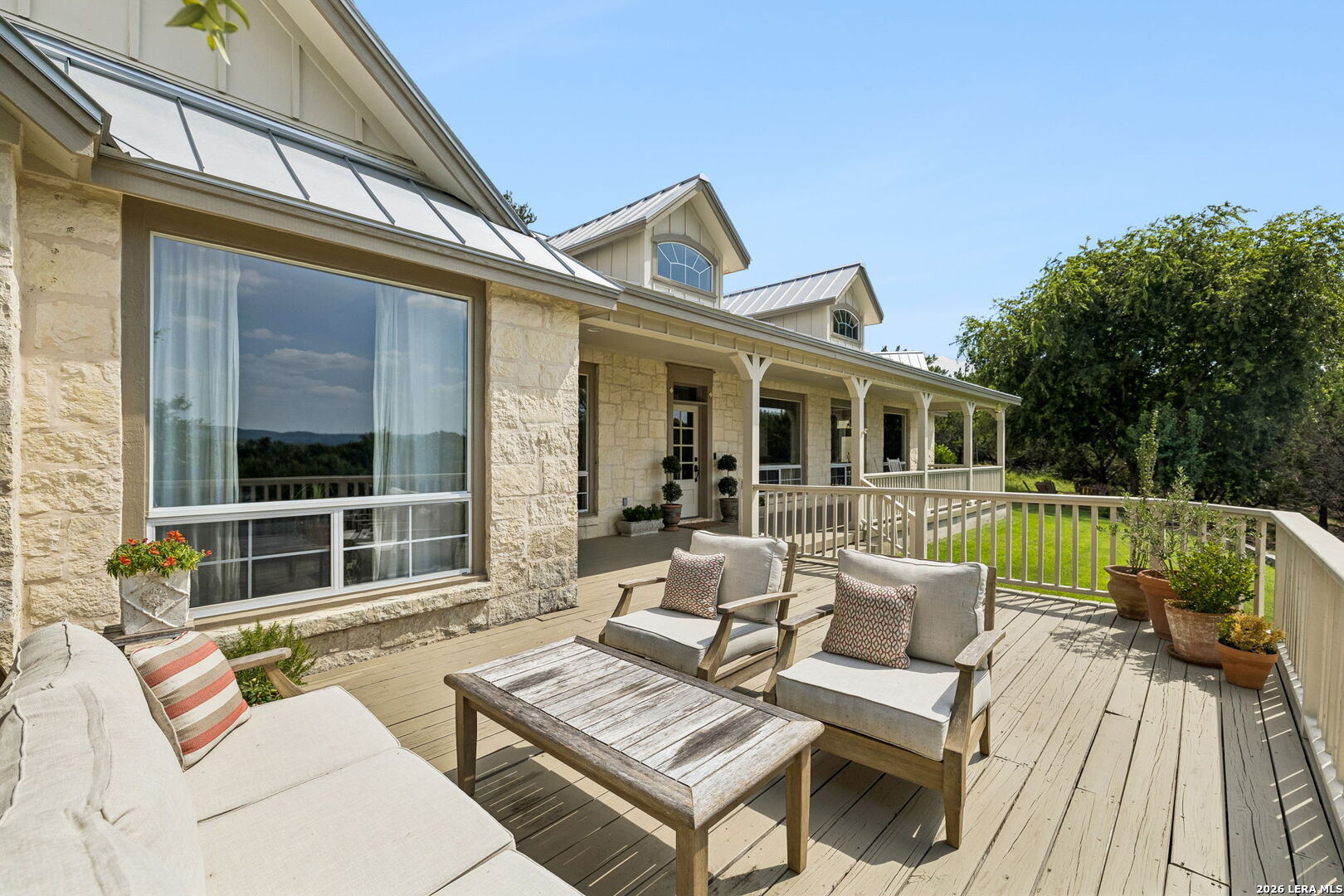 Furnished deck with cushioned seating beside a pale stone home and covered porch.