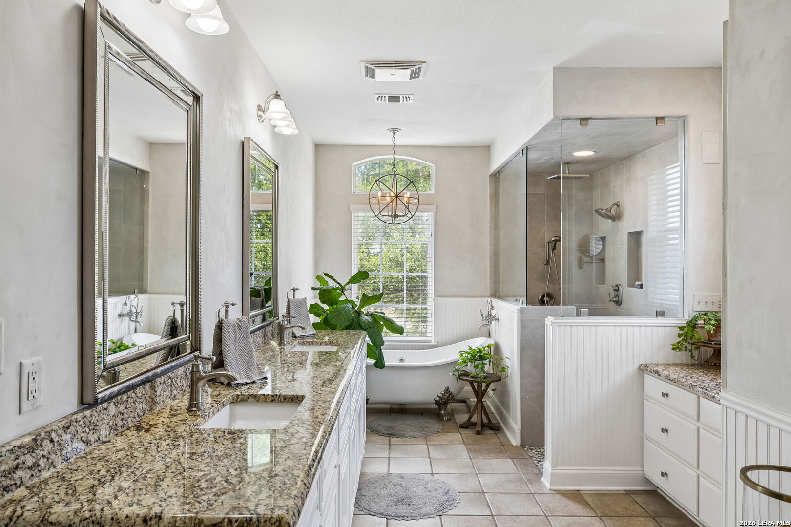 Bright bathroom with granite double vanity, freestanding tub beneath an arched window, and glass-enclosed shower.
