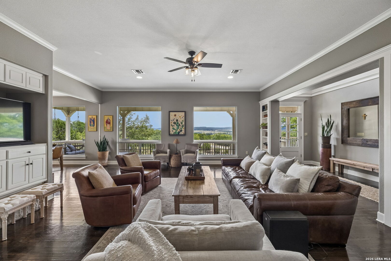 Furnished living room with leather seating and wide windows overlooking trees and distant hills.