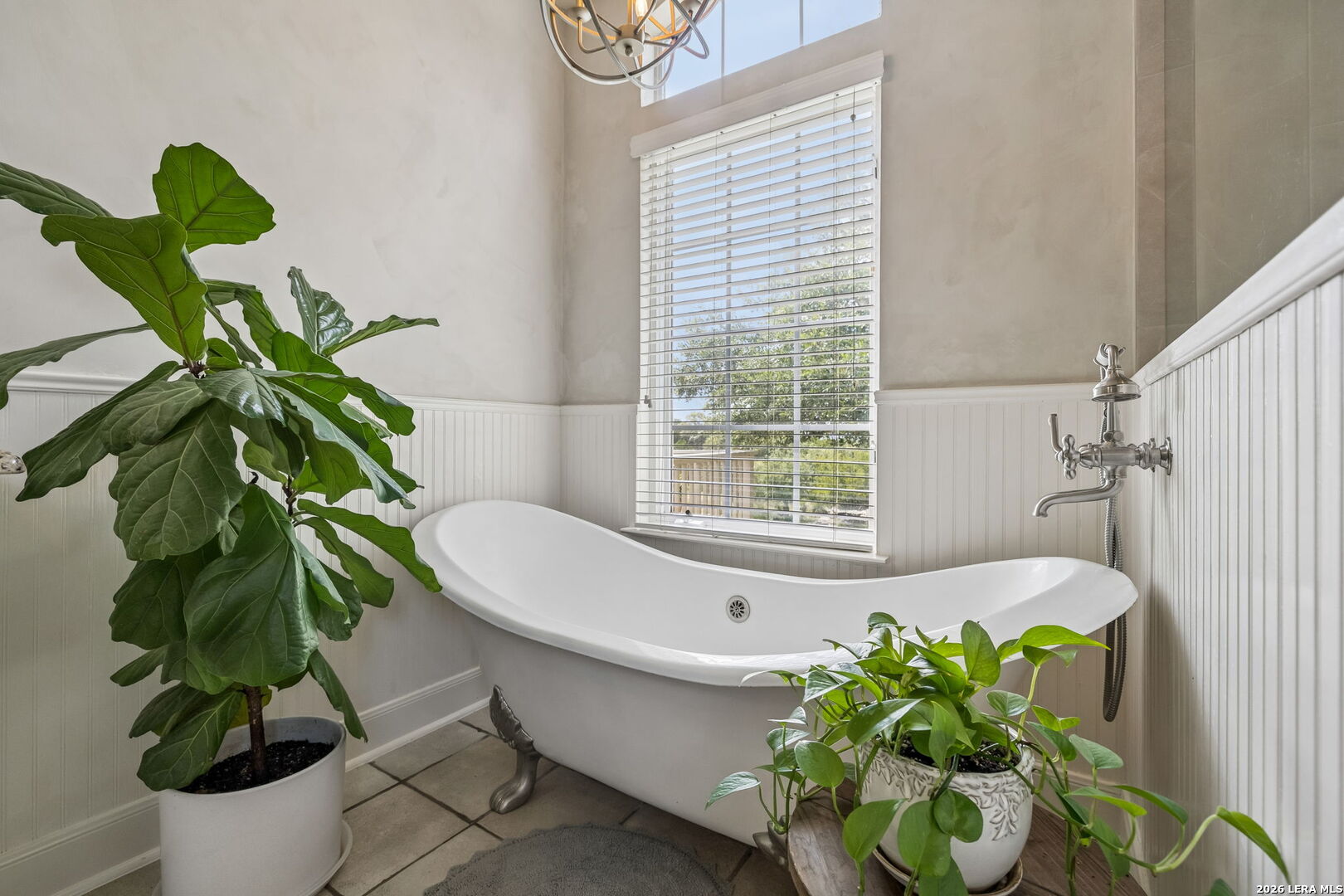 Bathroom with a freestanding clawfoot tub beneath a tall window, surrounded by leafy potted plants.