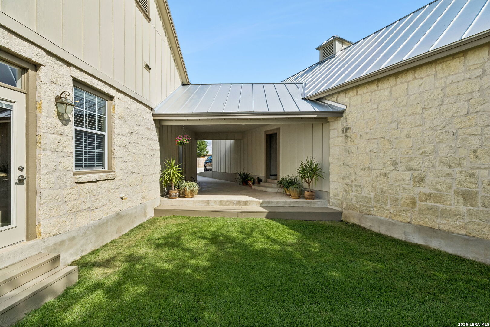 Stone house courtyard with a covered breezeway, potted plants, and a green lawn.