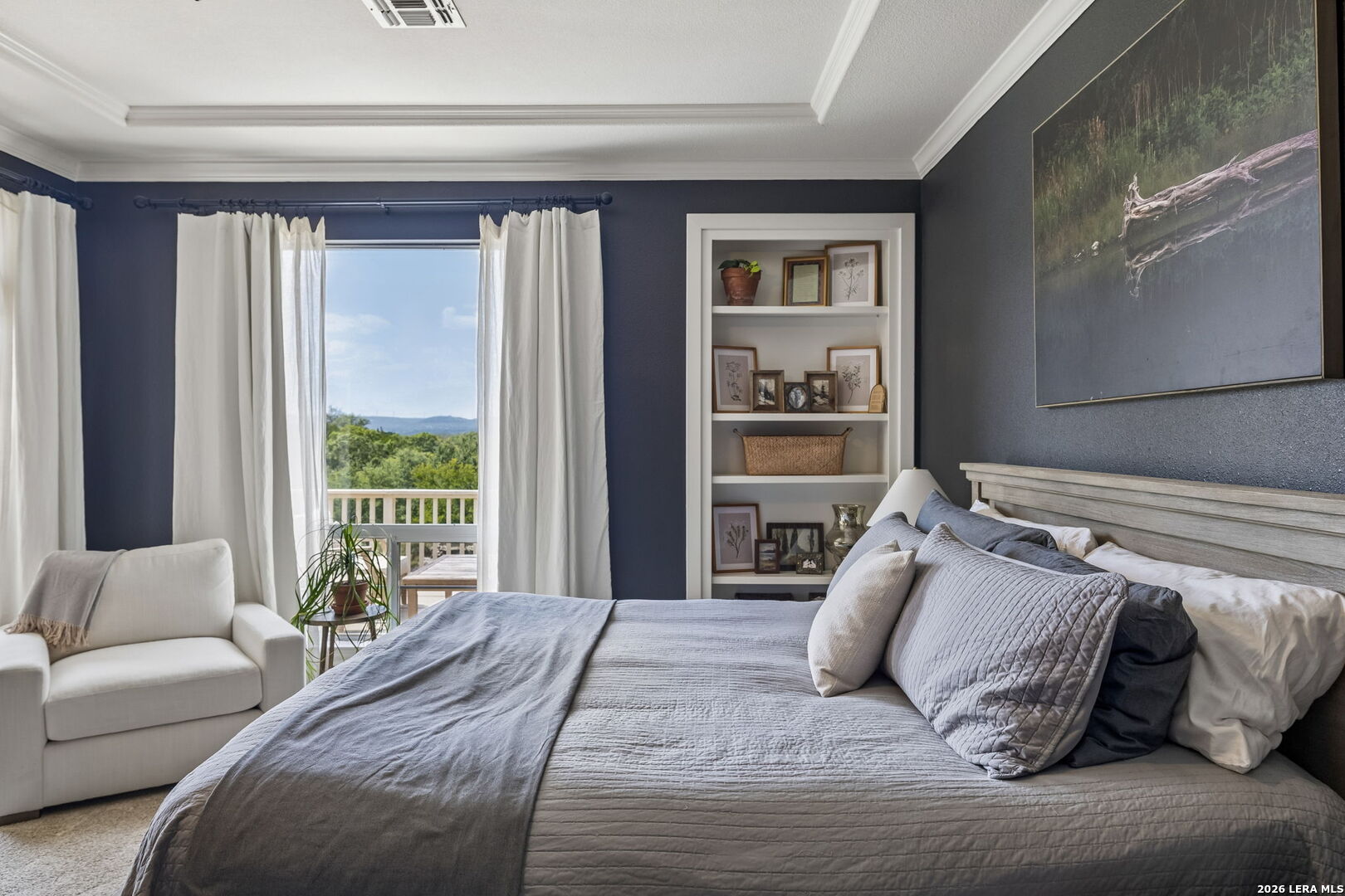 Bedroom with a gray quilted bed, white armchair, built-in shelves, and a balcony view of green trees.