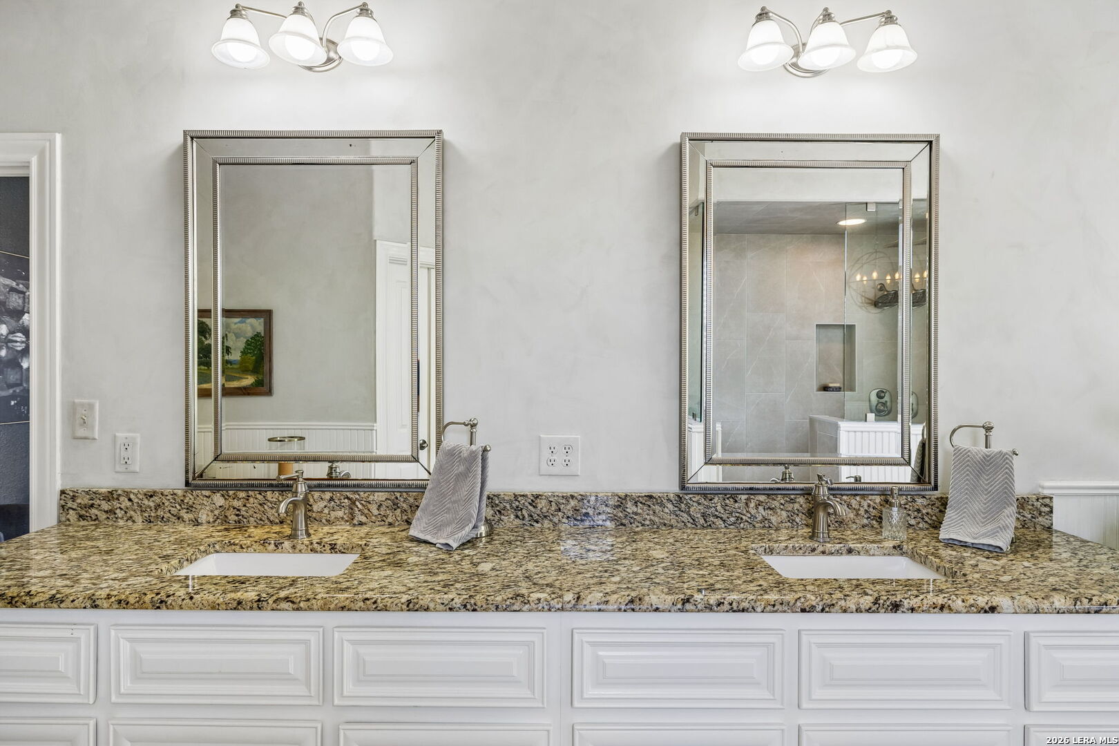 Bathroom vanity with double sinks, granite countertop, framed mirrors, and white paneled cabinetry.