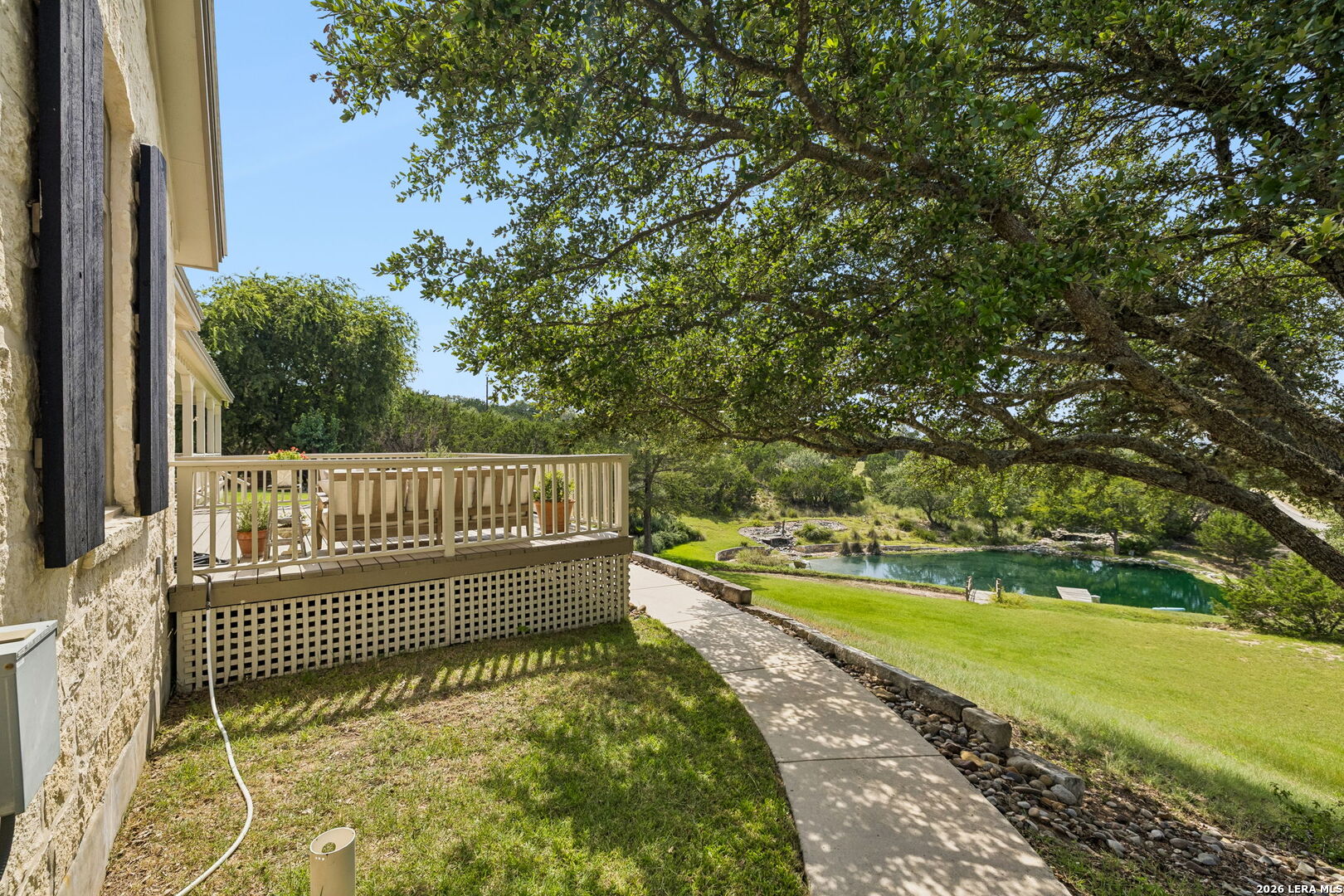 Stone home’s raised deck overlooks a green lawn and a small pool beneath a sprawling shade tree.
