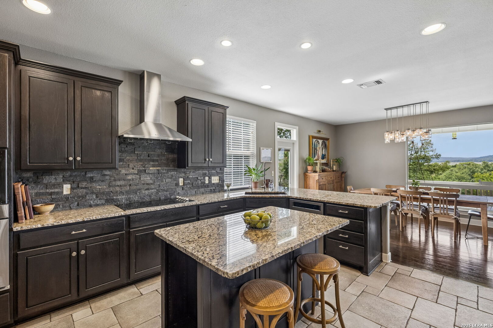 Kitchen with dark cabinets, granite island, stone backsplash, and dining area overlooking trees and distant hills.