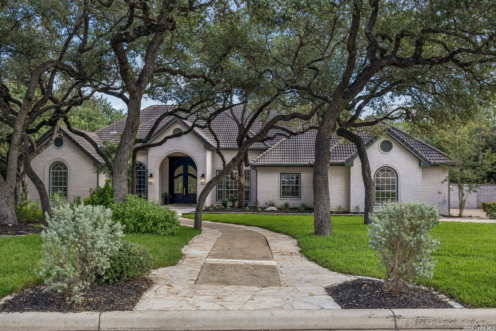 Light brick home with an arched entry, mature oak trees, and a stone walkway.