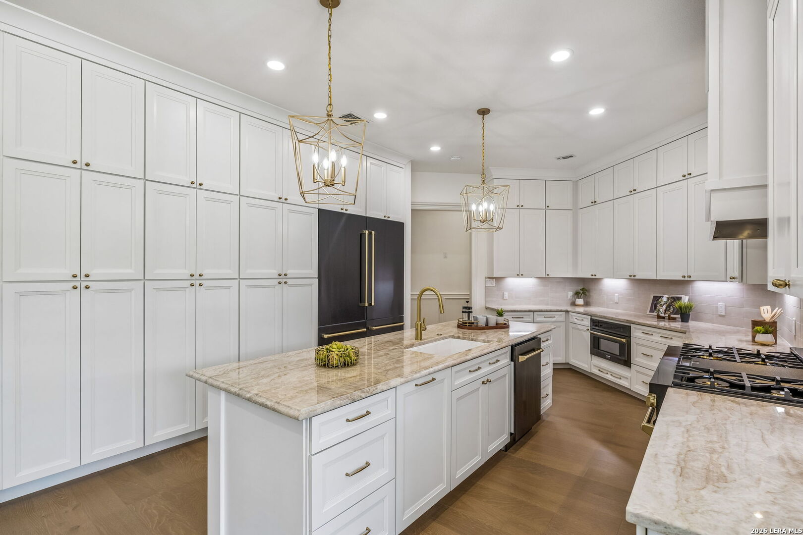 Bright kitchen with a large stone island, extensive white cabinetry, gold fixtures, and a dark refrigerator.