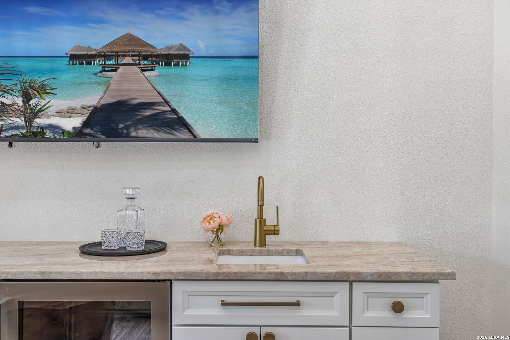 Wet bar with a stone counter, brass faucet, white cabinetry, beverage refrigerator, and wall-mounted television.