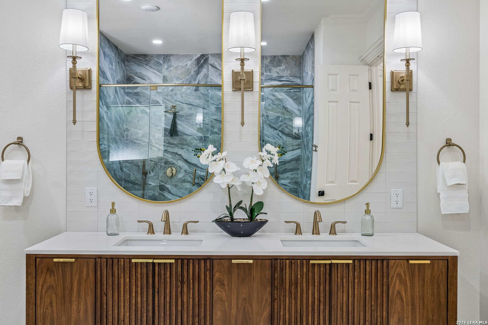 Bathroom with a double-sink wood vanity, brass fixtures, and gold-framed mirrors reflecting a blue-gray marble shower.