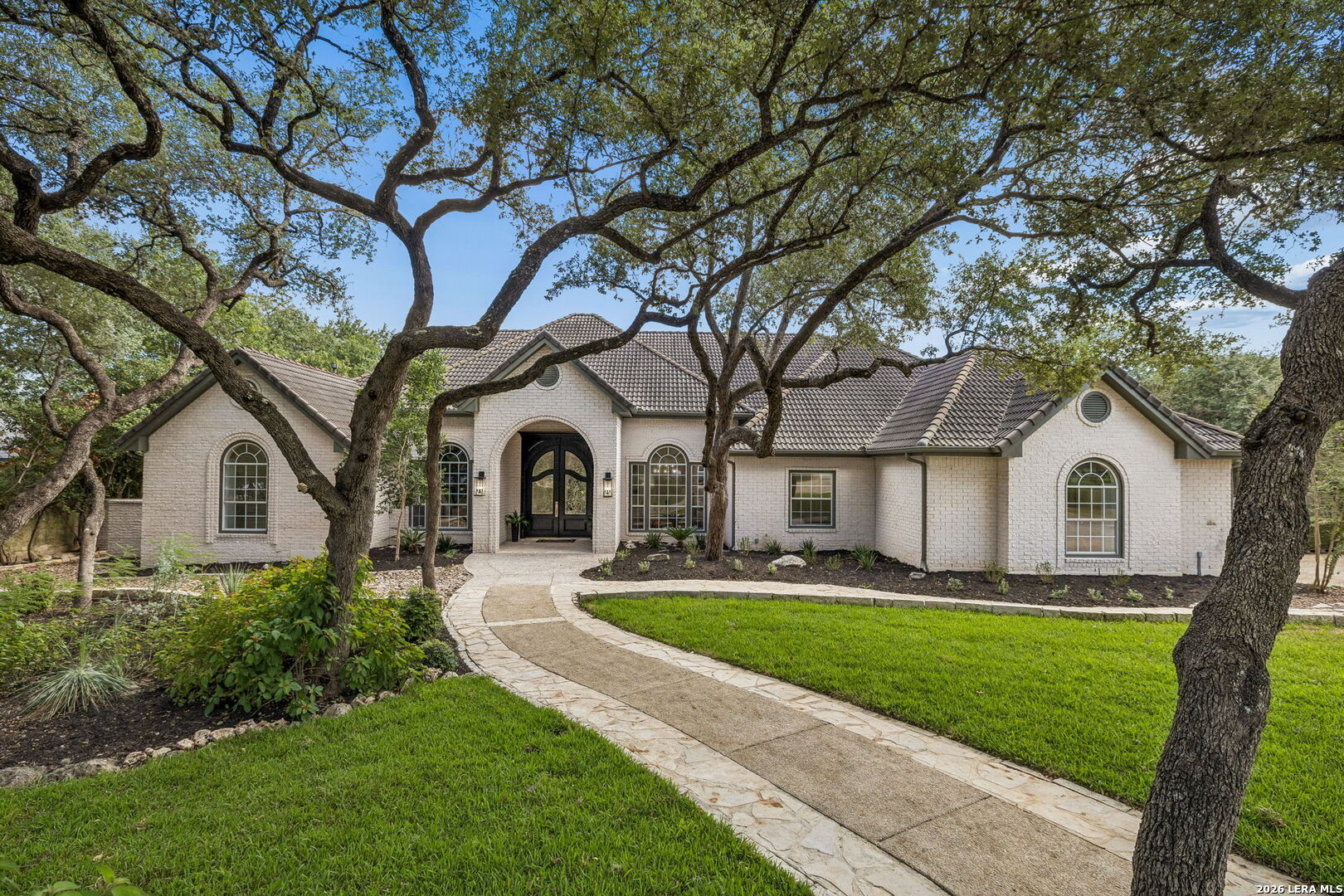 White brick home framed by mature oak trees, with a curved stone walkway to the arched entry.