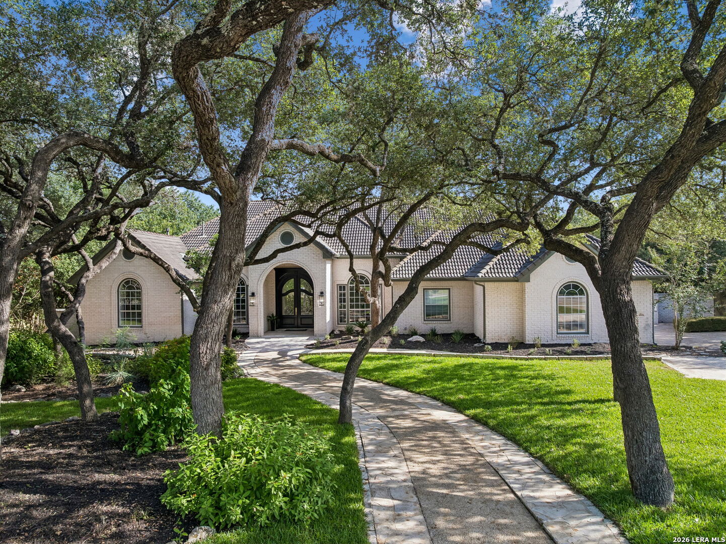 Light brick home with a tiled roof, framed by mature oak trees and a curved stone walkway.