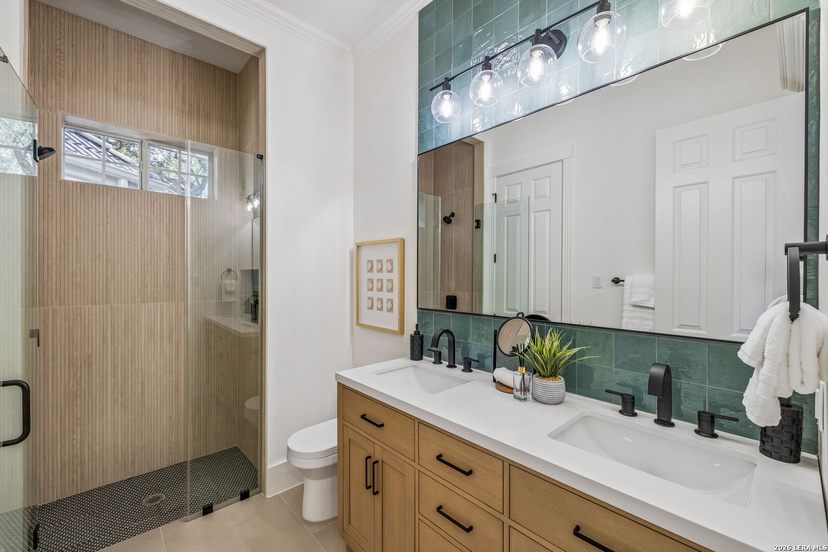 Bathroom with an oak double vanity, green tile backsplash, and glass-enclosed shower with beige vertical tile.