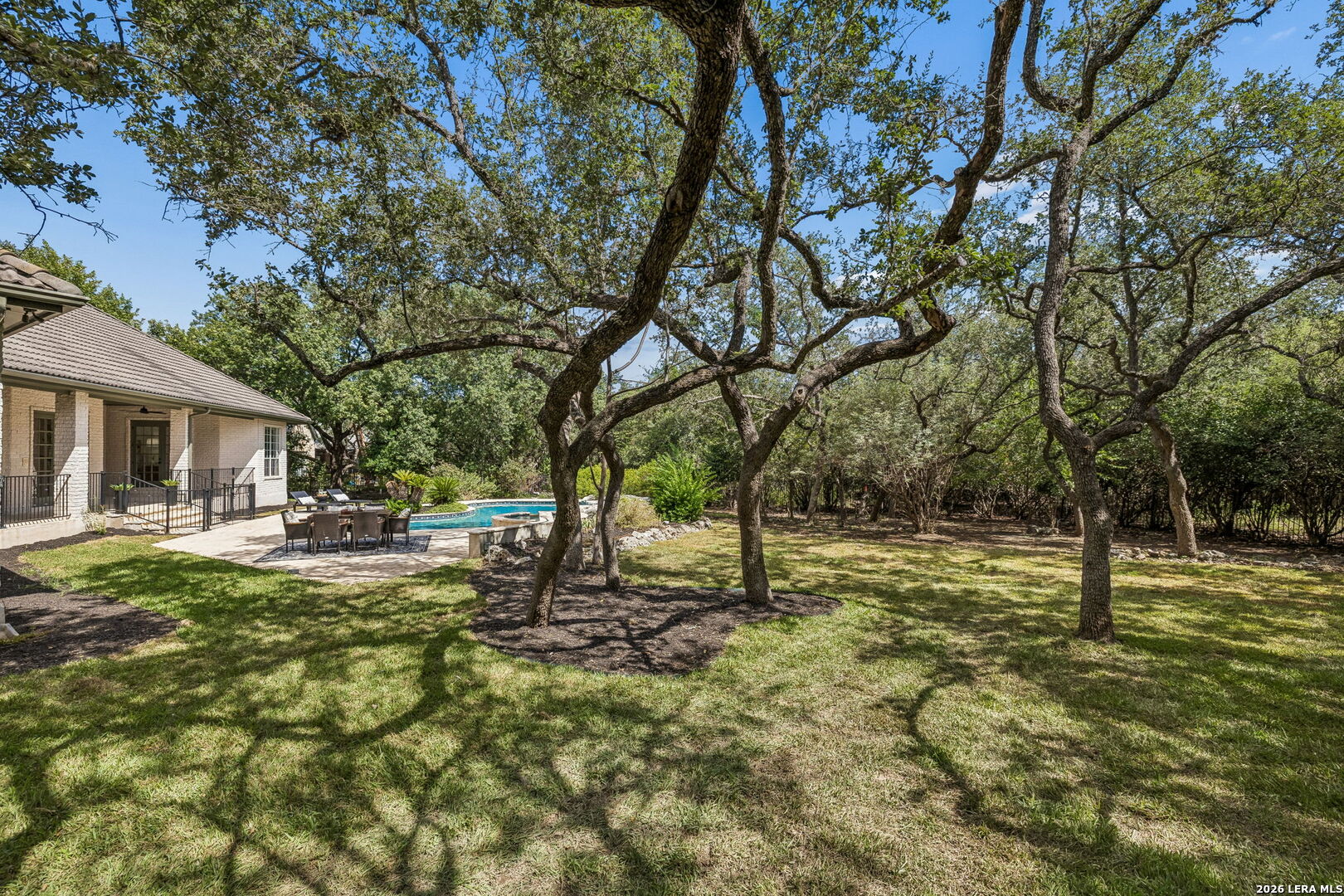 Shaded backyard with mature oak trees, a swimming pool, and a stone patio with outdoor dining furniture.