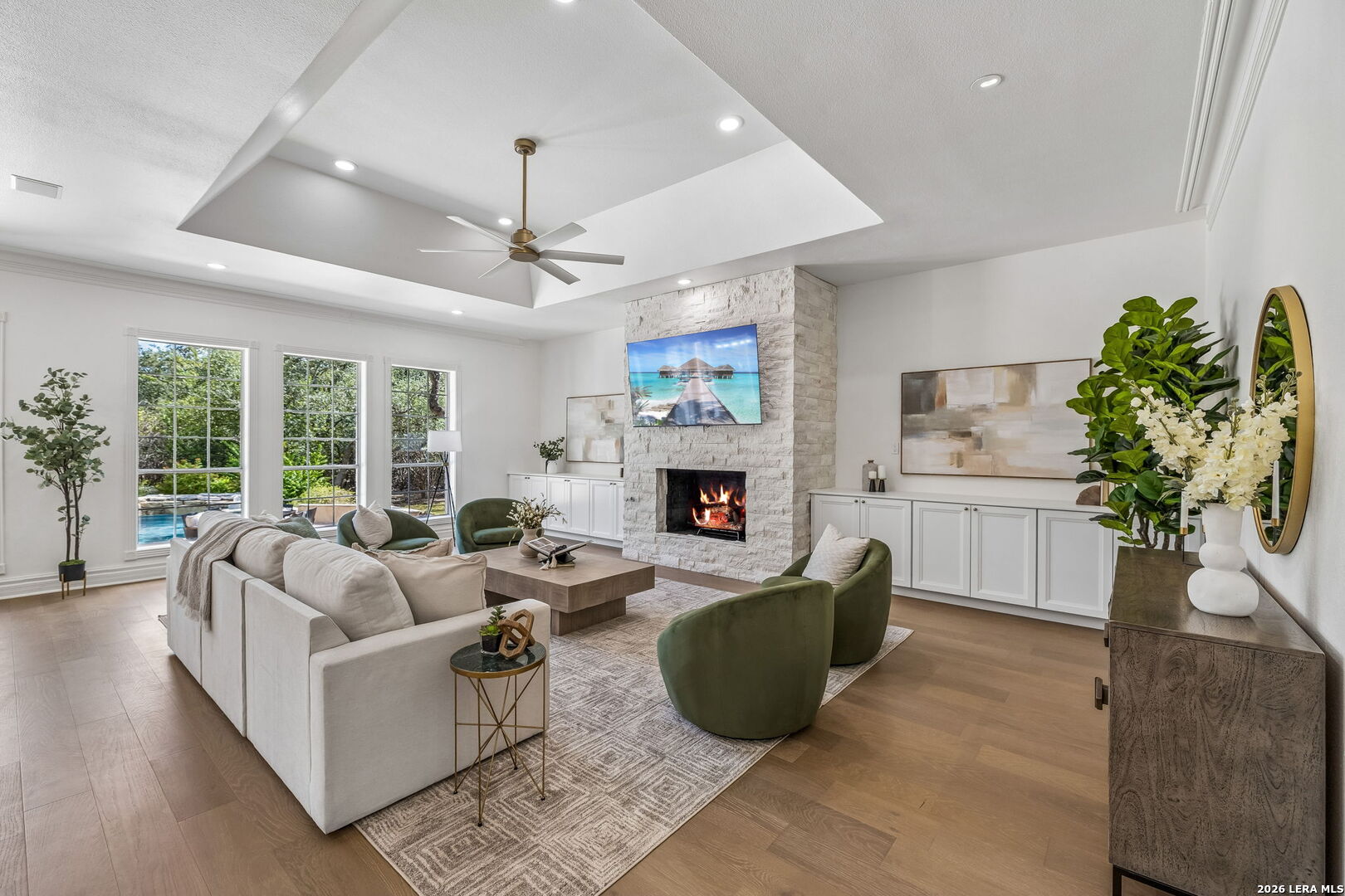 Living room with a white stone fireplace, tray ceiling, and tall windows overlooking a pool.