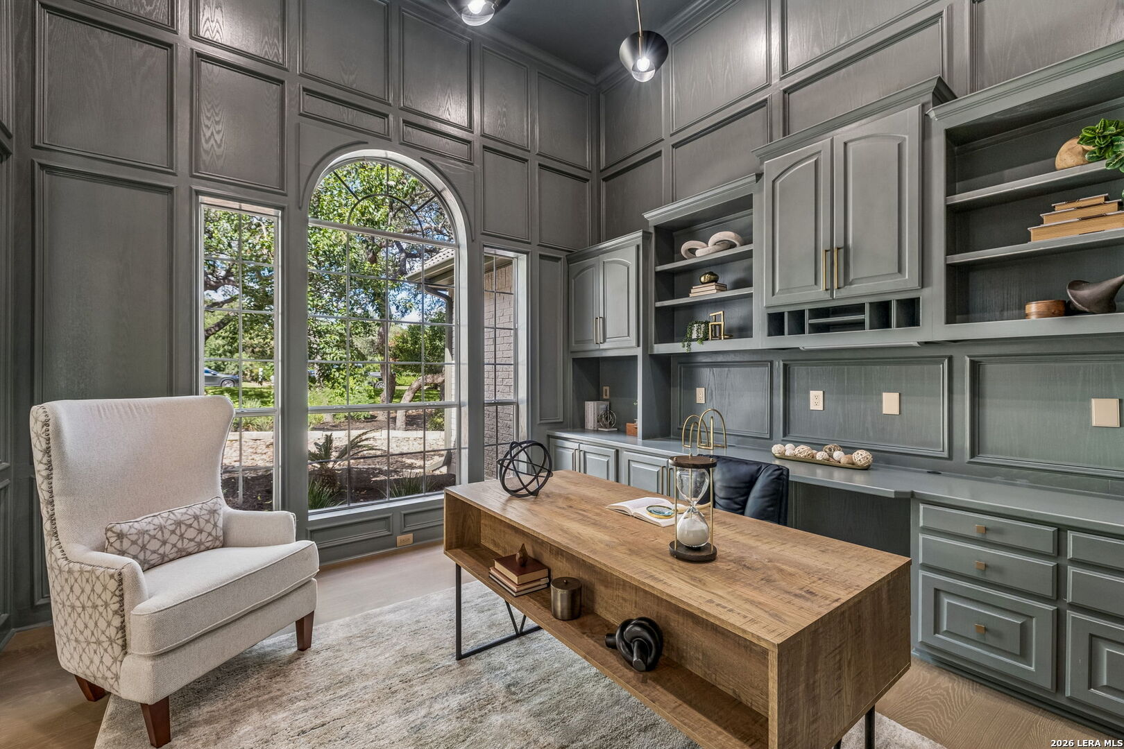 Furnished home office with dark paneled walls, built-in shelving, and a large arched window.