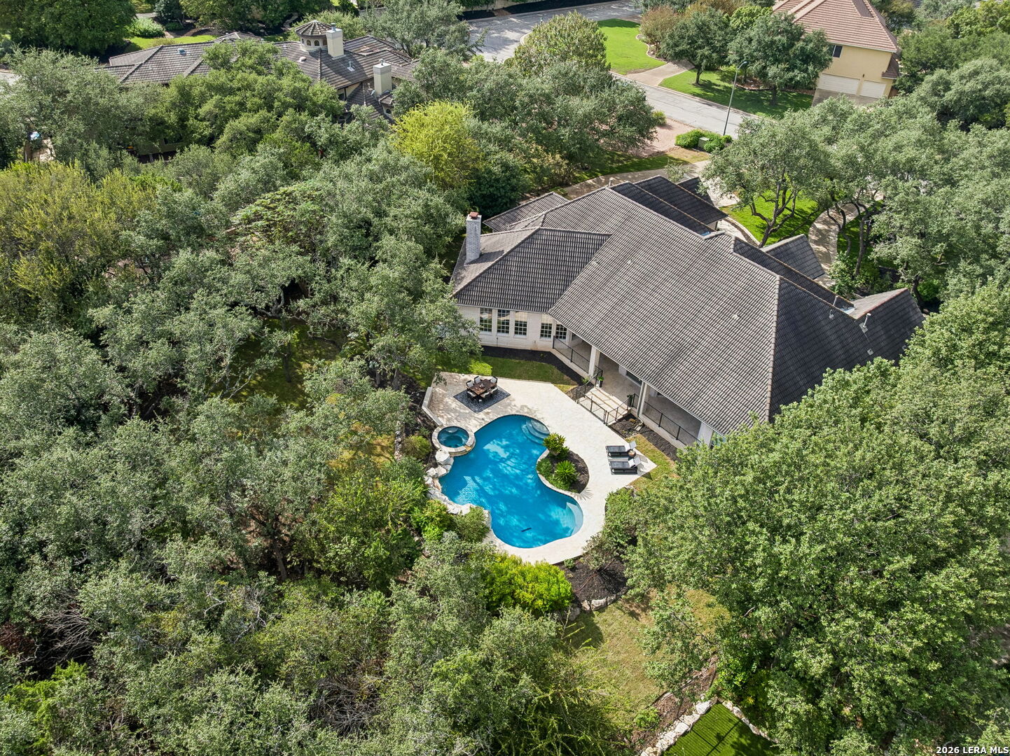 Aerial view of a tiled-roof home with a backyard pool and spa amid mature trees.