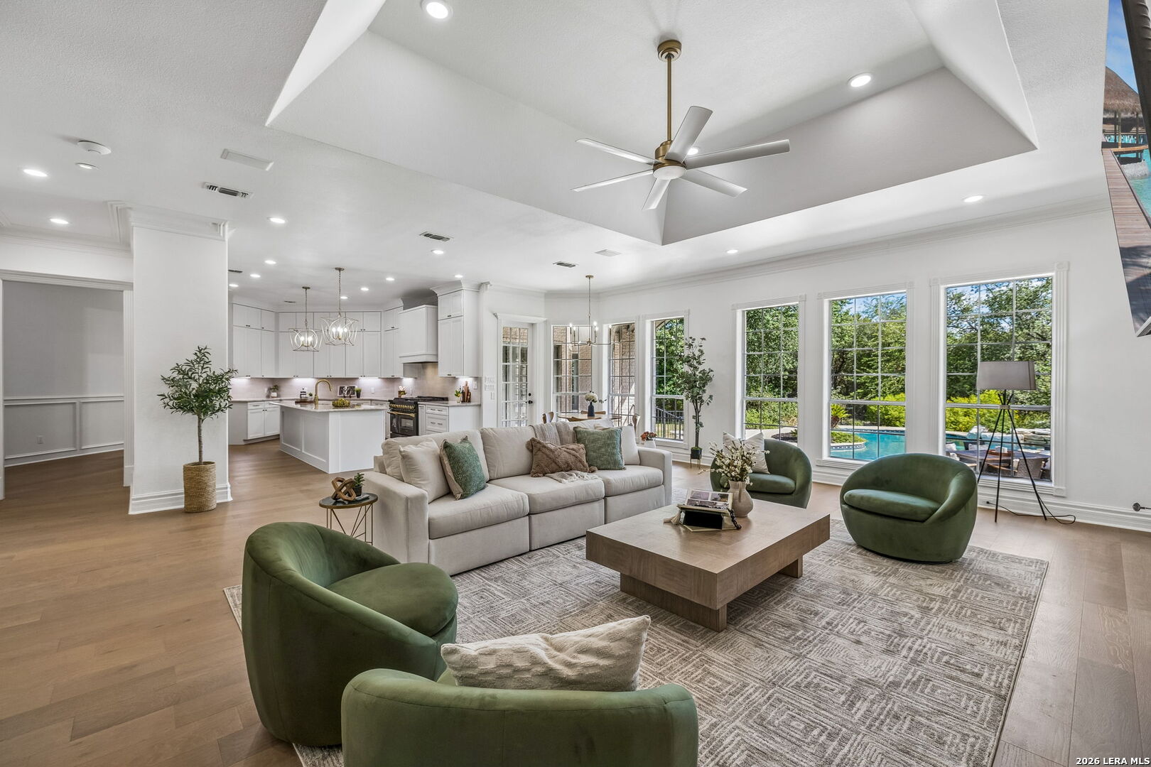 Open-plan living room with green lounge chairs, white kitchen, and pool views through large windows.