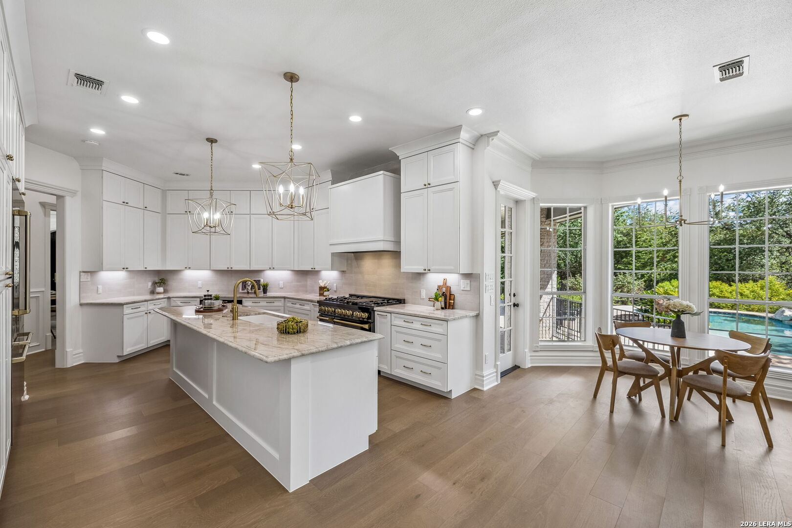 Bright white kitchen with a marble island and dining area beside windows overlooking a pool.