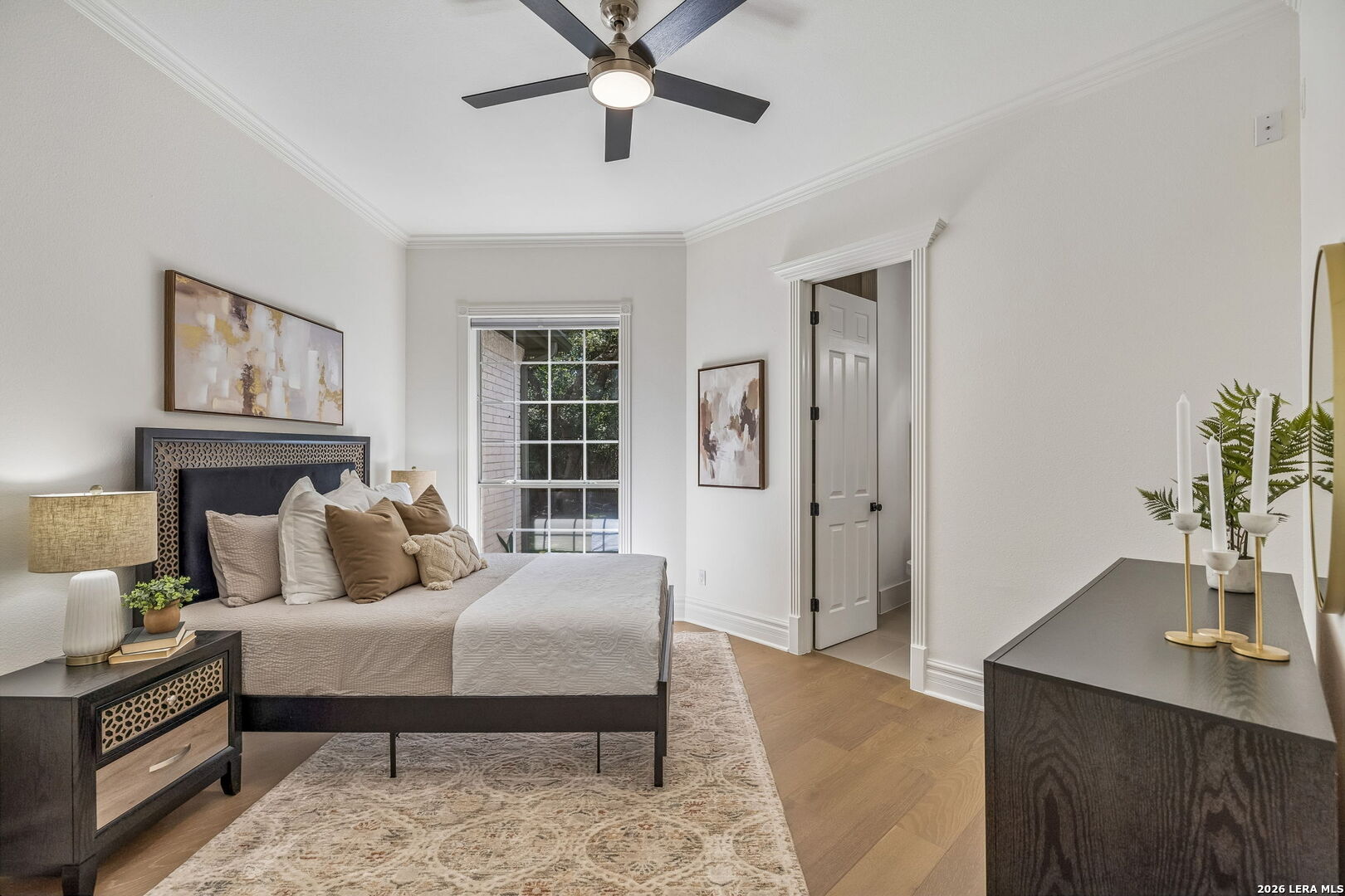 Staged bedroom with a dark bed, neutral bedding, ceiling fan, and large window.