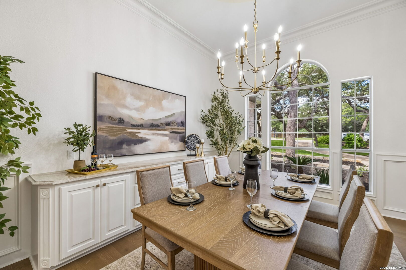 Staged dining room with a set wooden table, white sideboard, brass chandelier, and tall arched windows.