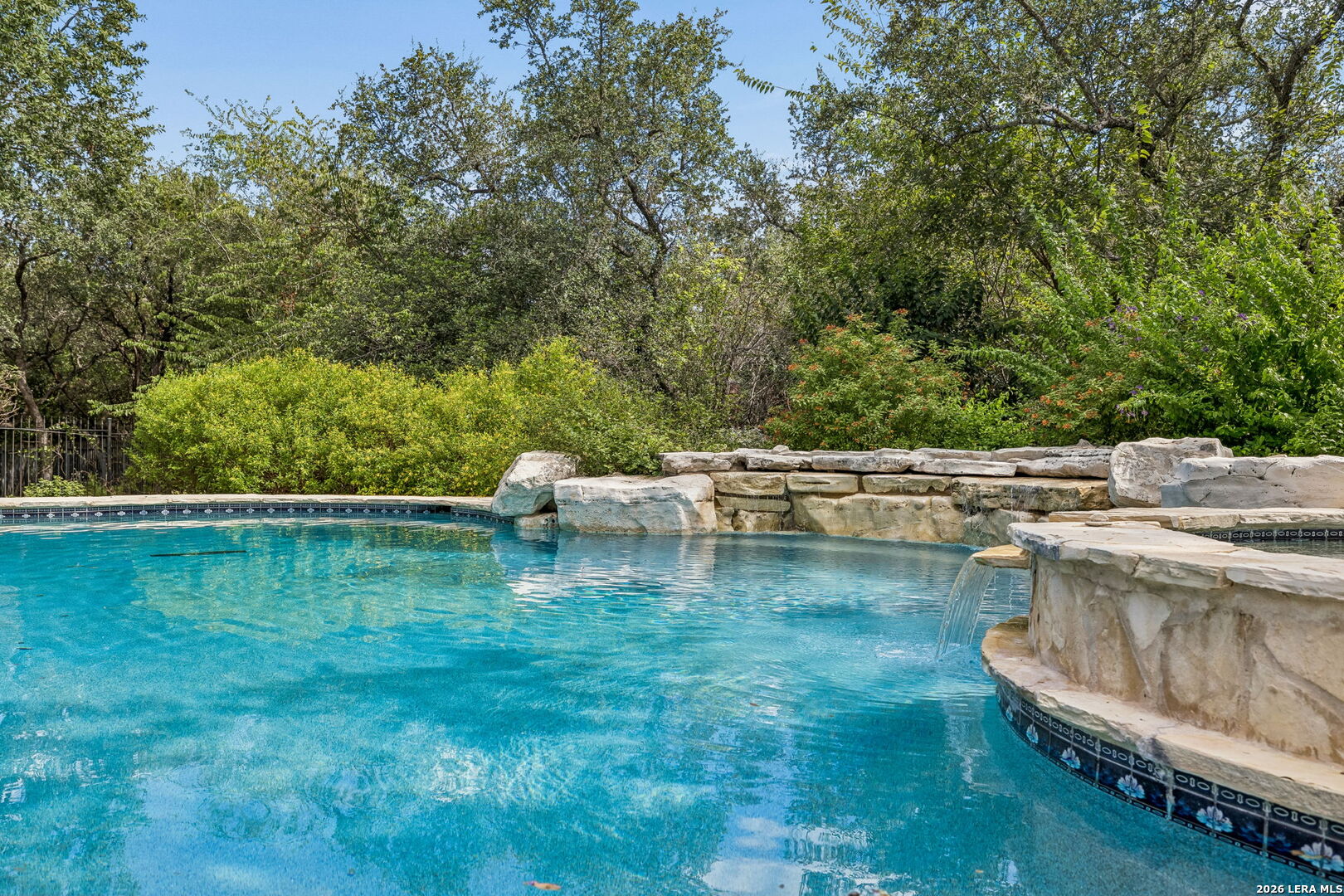 Outdoor pool with a natural stone wall, small waterfall, and mature trees.
