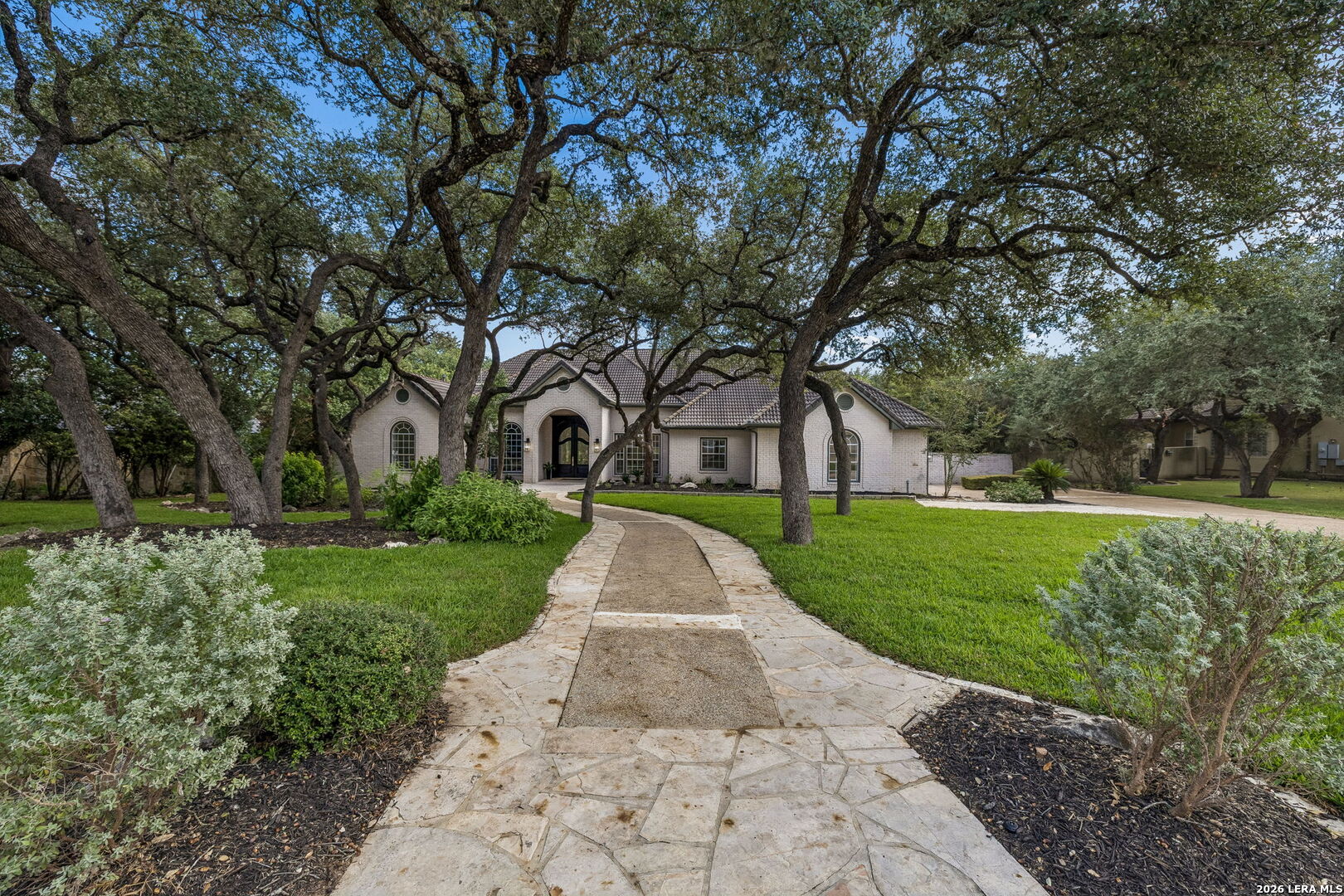Light brick home with an arched entrance, approached by a curved stone walkway beneath mature oak trees.
