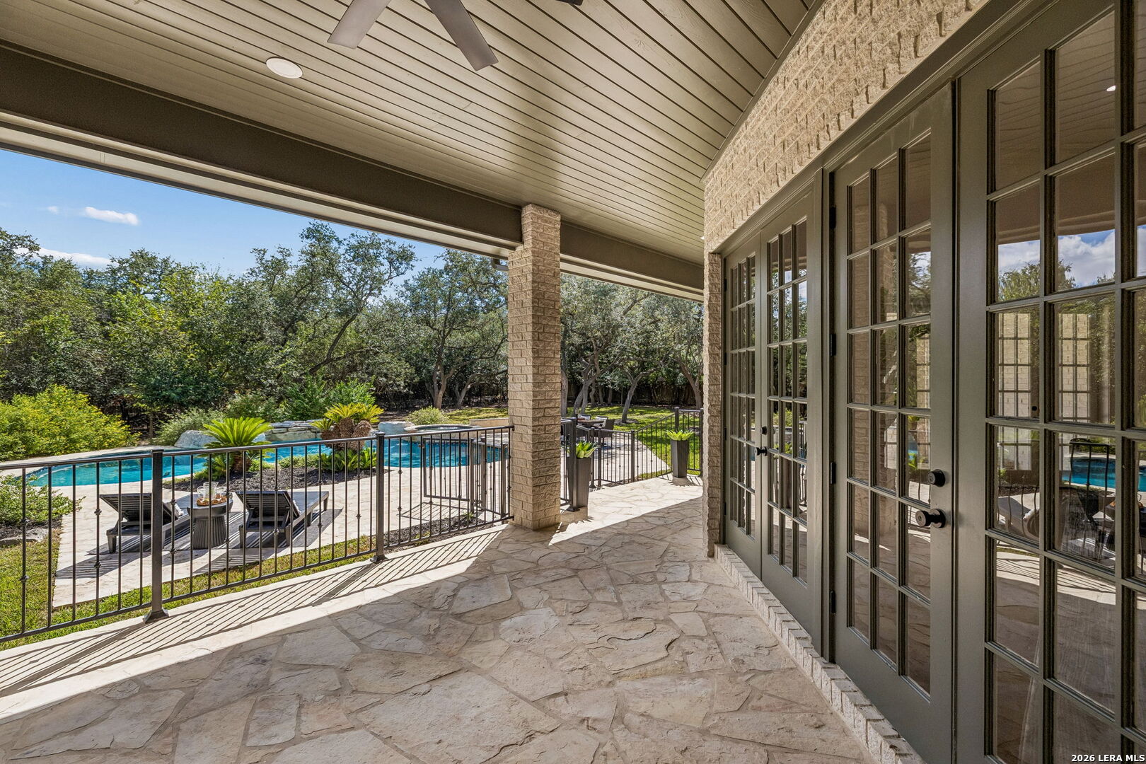 Covered stone patio overlooking a pool and leafy backyard, with French doors and a ceiling fan.