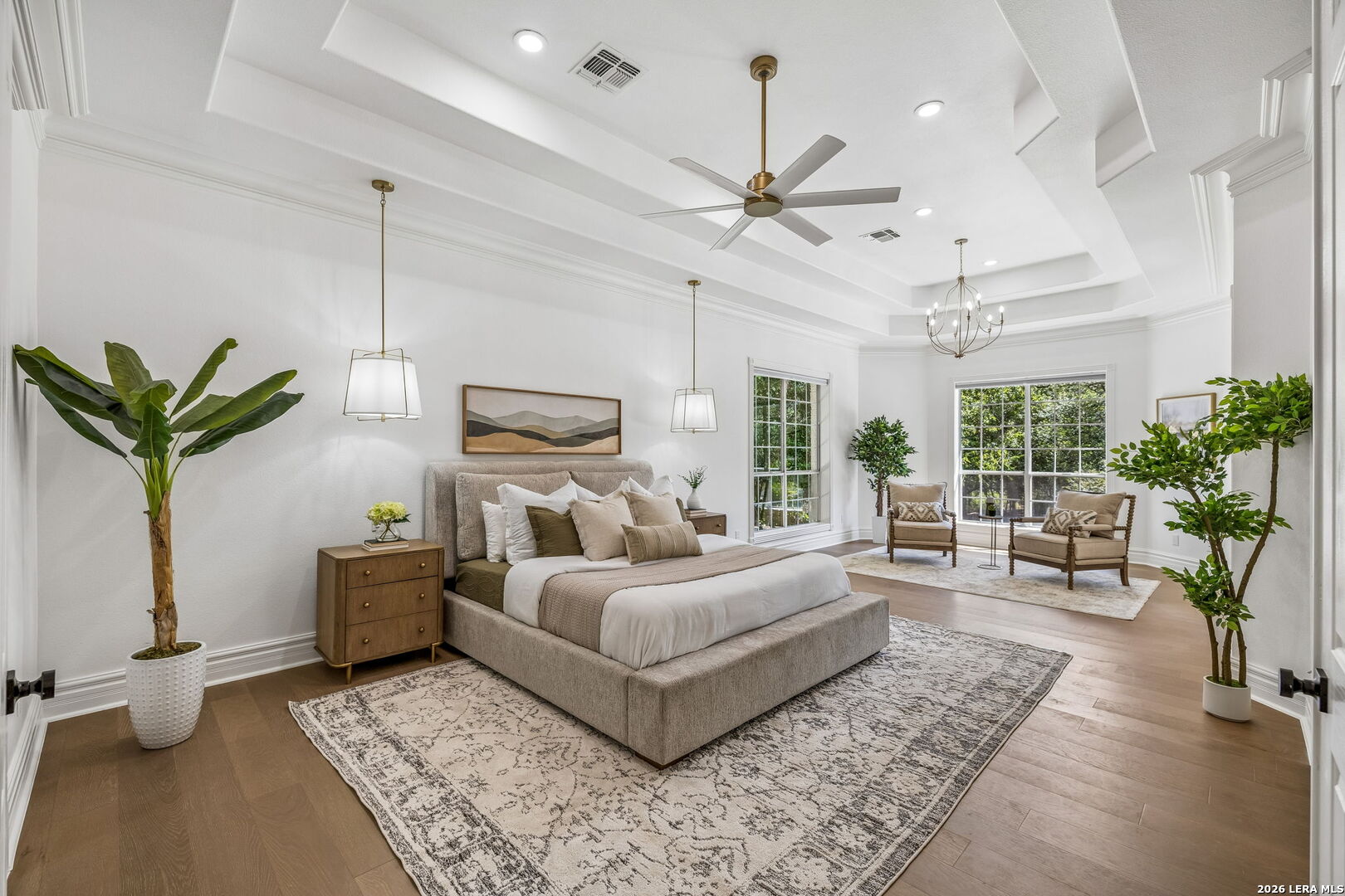 Staged bedroom with a large upholstered bed, tray ceiling, and bright window-side sitting area.