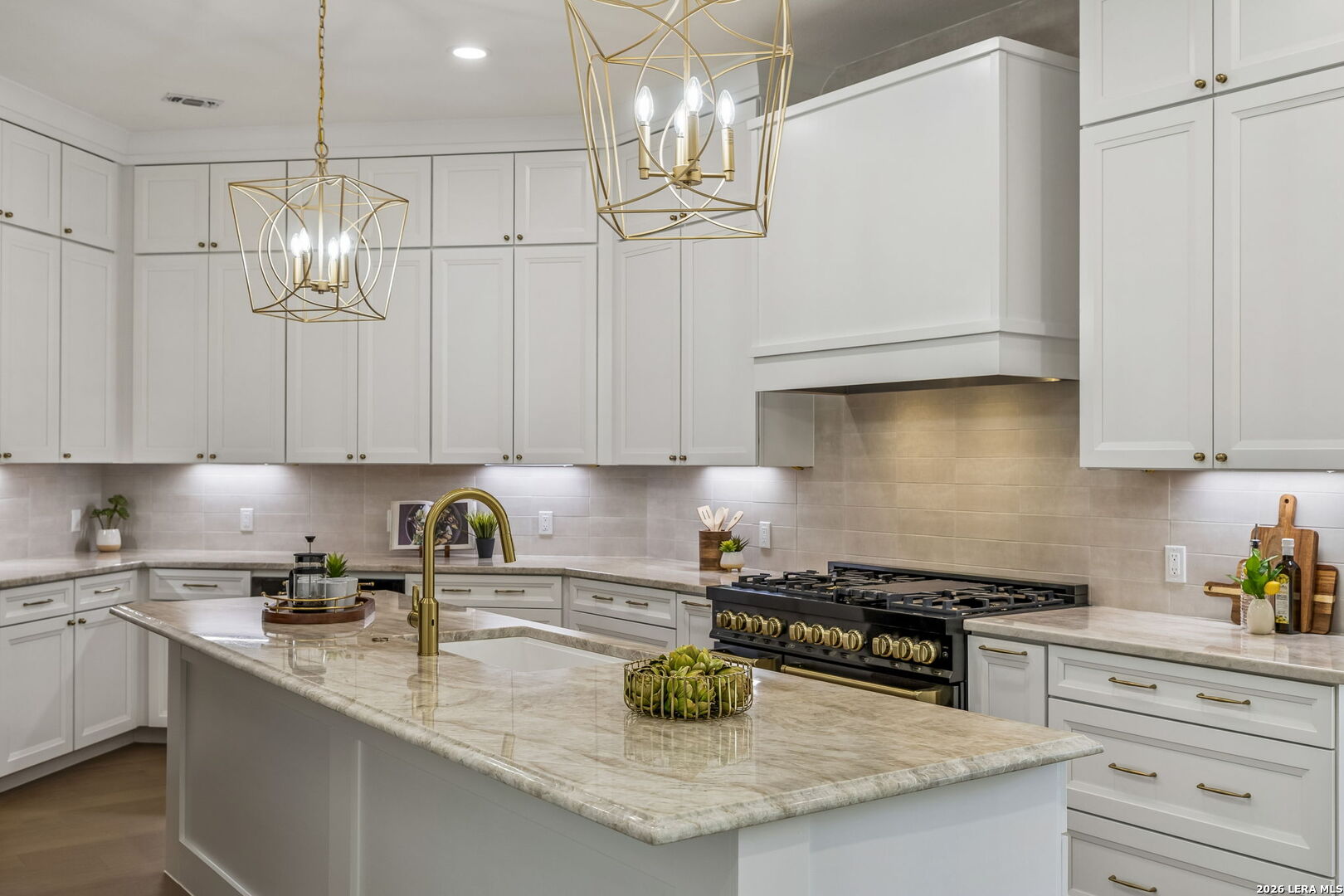 Bright white kitchen with a large marble-patterned island, brass fixtures, and a wide black gas range.