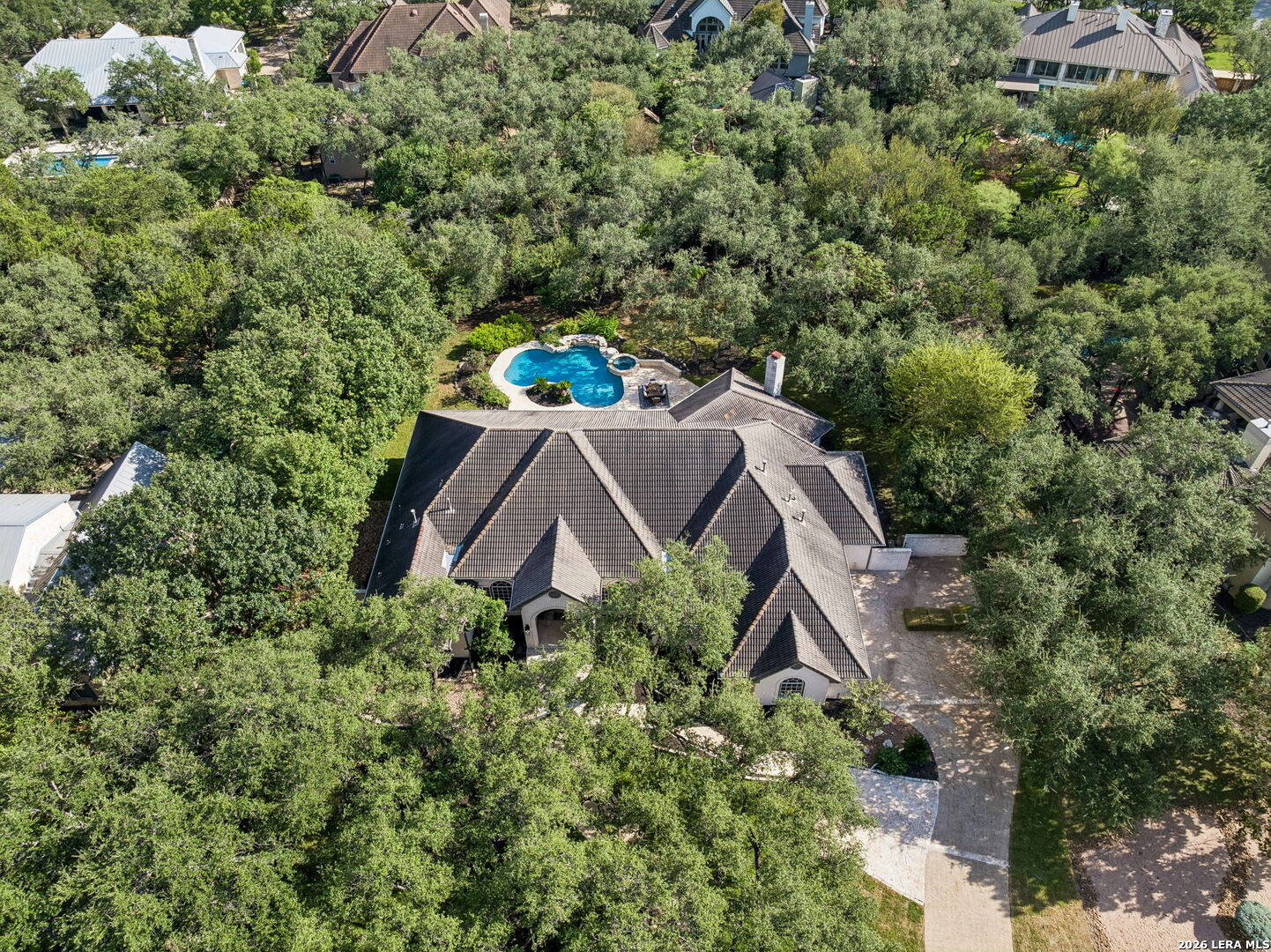 Aerial view of a tree-surrounded home with a swimming pool and broad stone driveway.