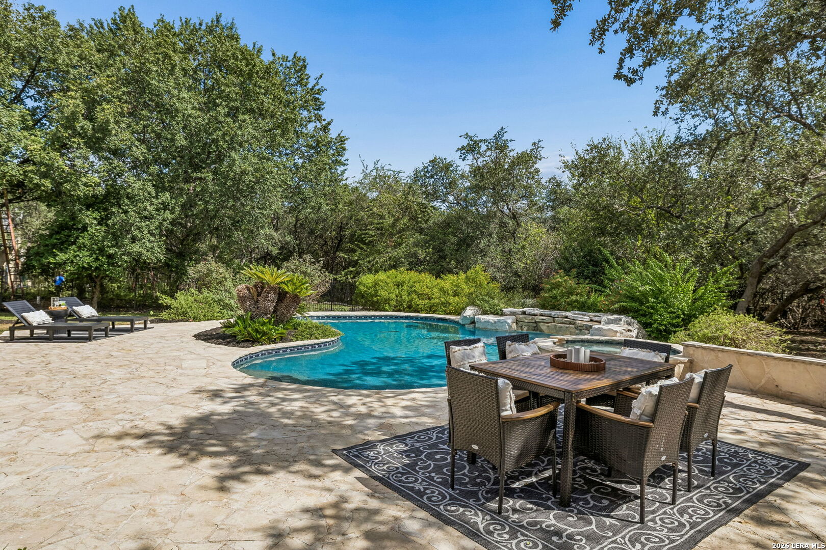 Backyard pool bordered by stone patio, with outdoor dining furniture and lounge chairs.