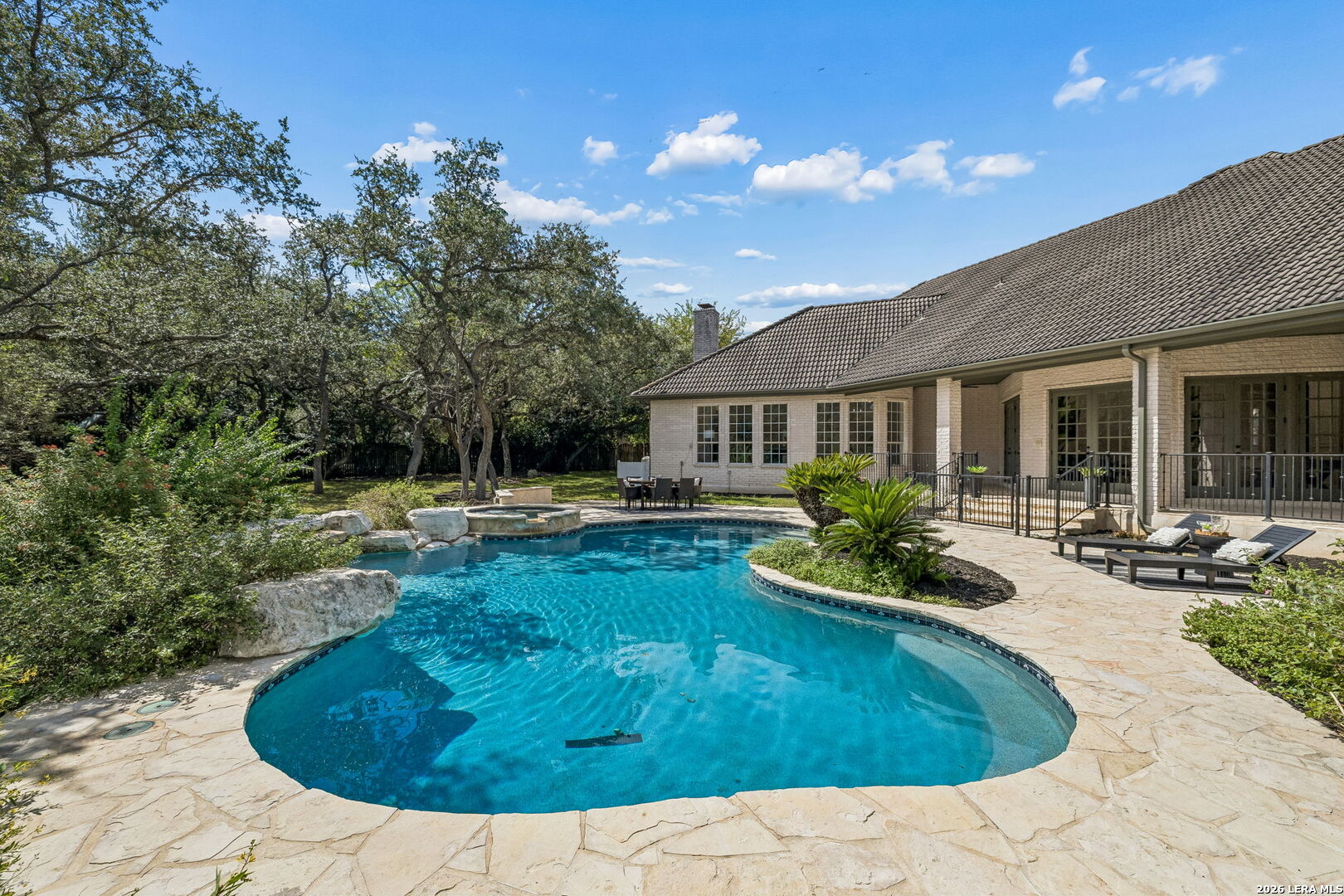 Backyard swimming pool with a curved edge, stone patio, lounge seating, and mature trees.