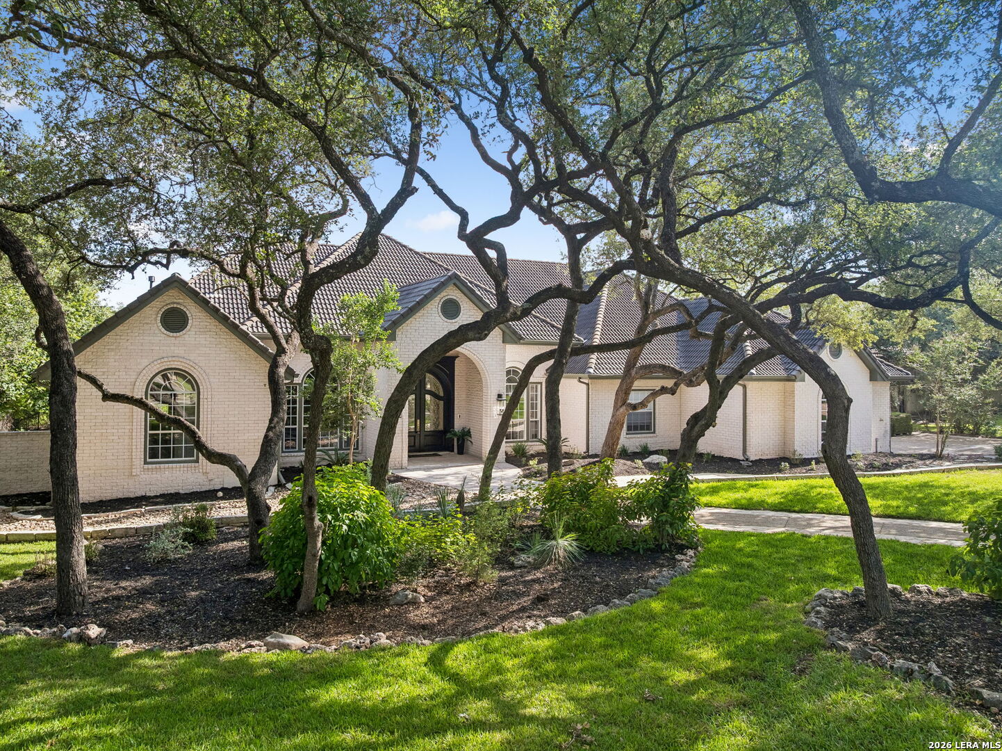 White brick home with an arched entry, tile roof, and mature oak trees across the front lawn.
