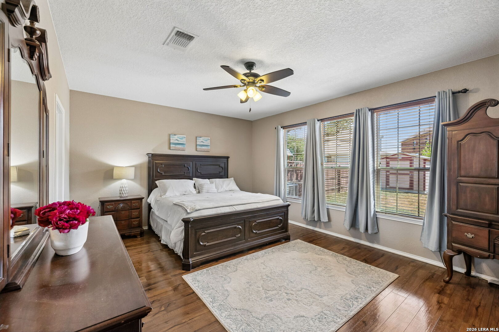 Furnished bedroom with a dark wood bed, hardwood floors, and three curtained windows.