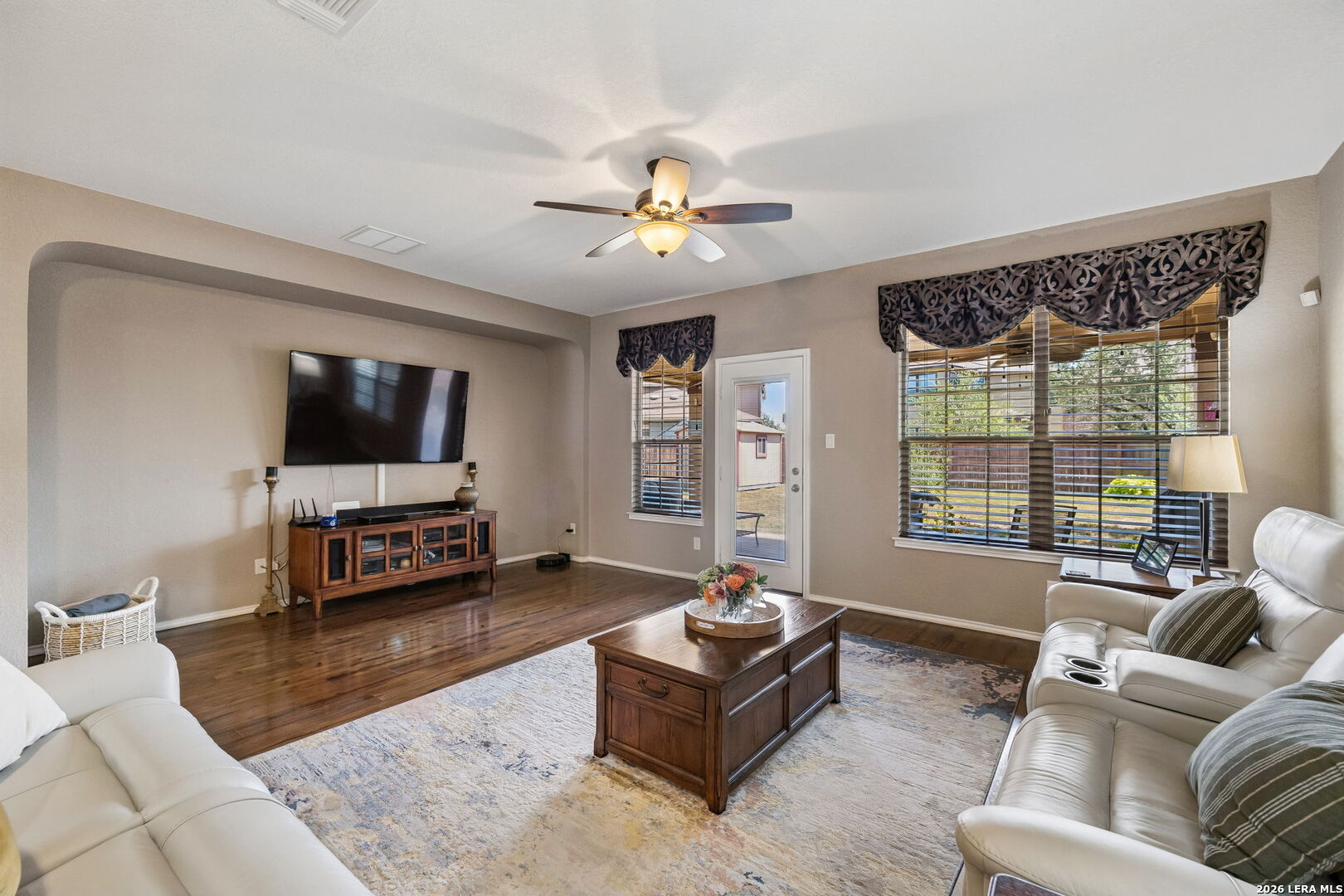 Living room with white leather sofas, hardwood floors, and large windows overlooking the backyard.