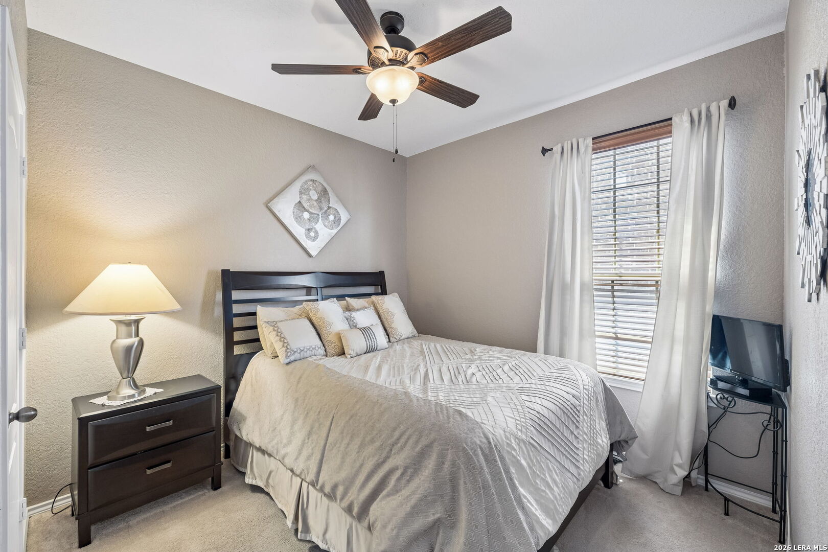 Furnished bedroom with pale bedding, a dark wood bed, ceiling fan, and curtained window.