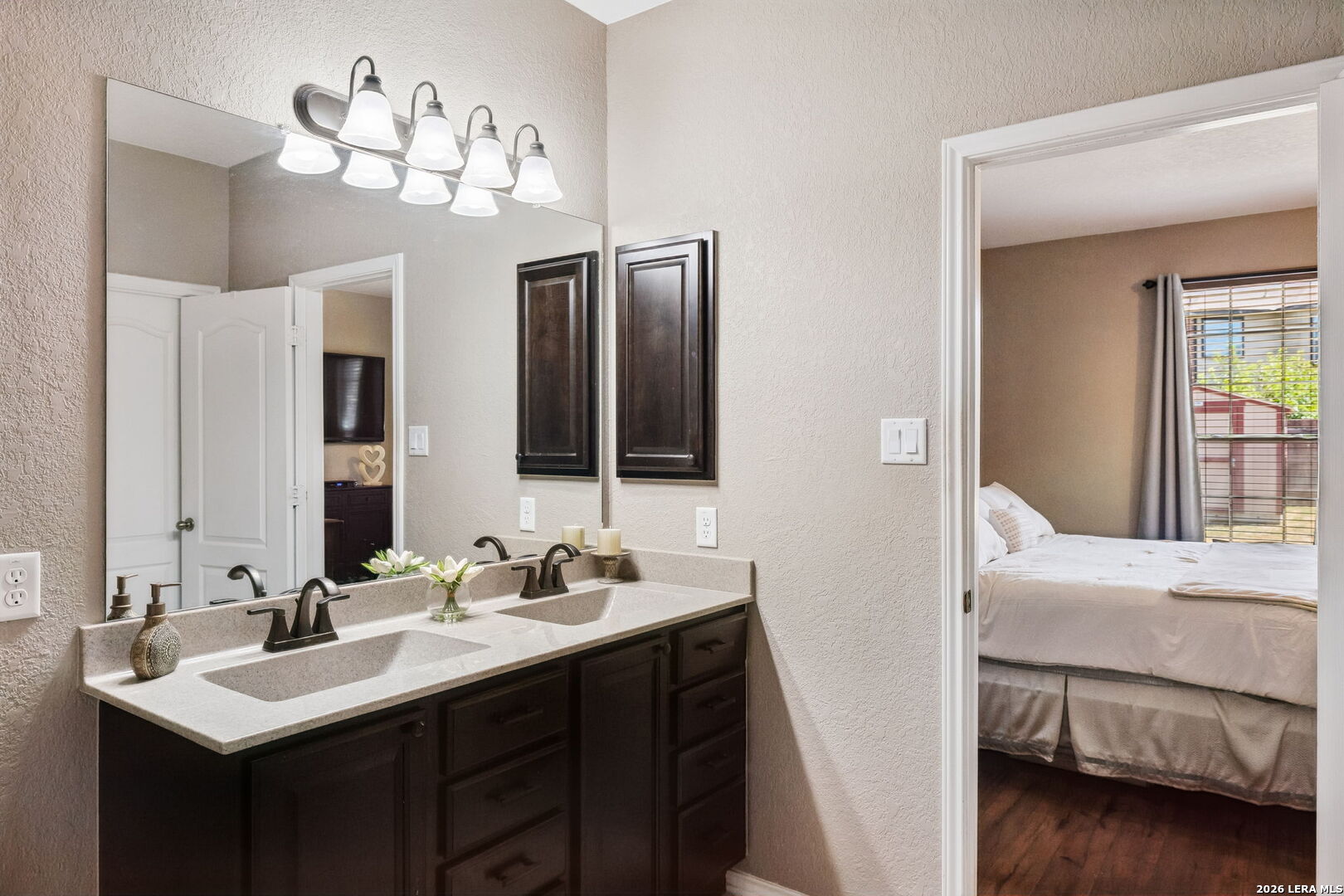Bathroom double vanity with dark cabinetry and a doorway to a bedroom with a window.