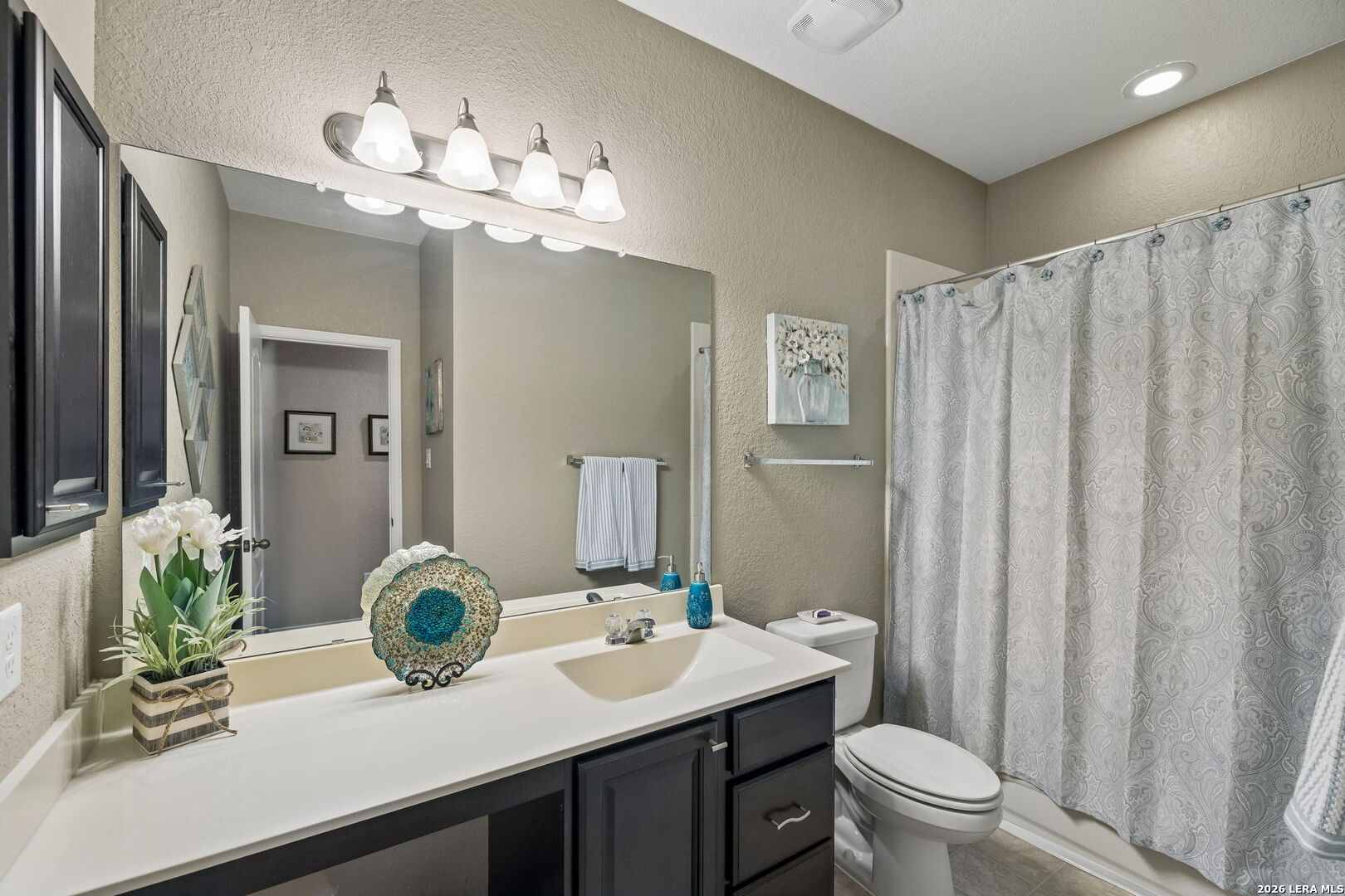 Bathroom with a long white vanity, dark cabinets, large mirror, and patterned gray shower curtain.