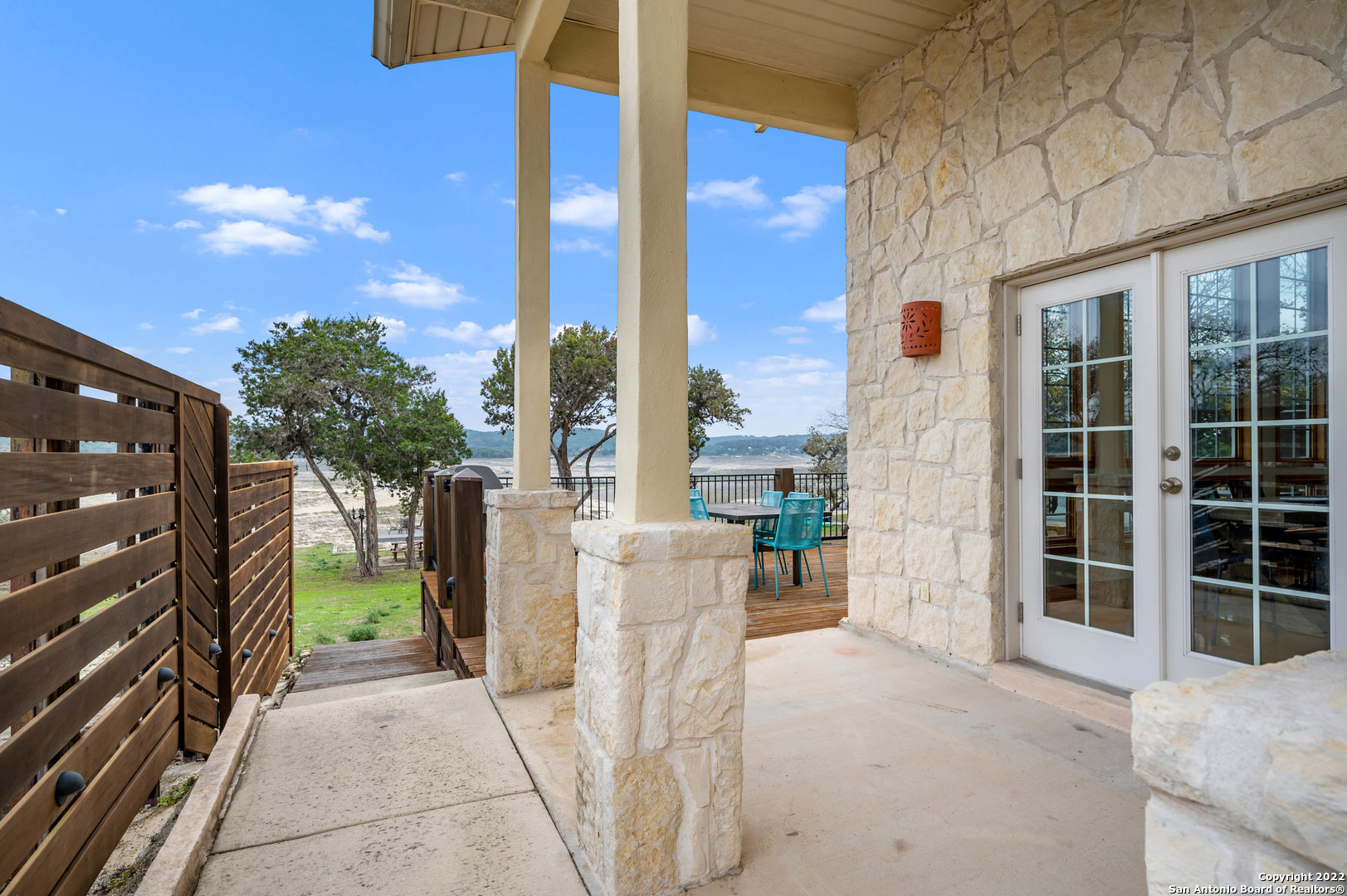 Covered stone porch overlooking a lake, with a wooden deck and turquoise patio furniture.