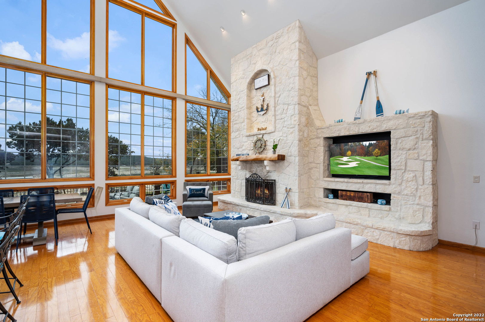 Living room with a stone fireplace, white sectional, polished wood floors, and tall wood-framed windows.