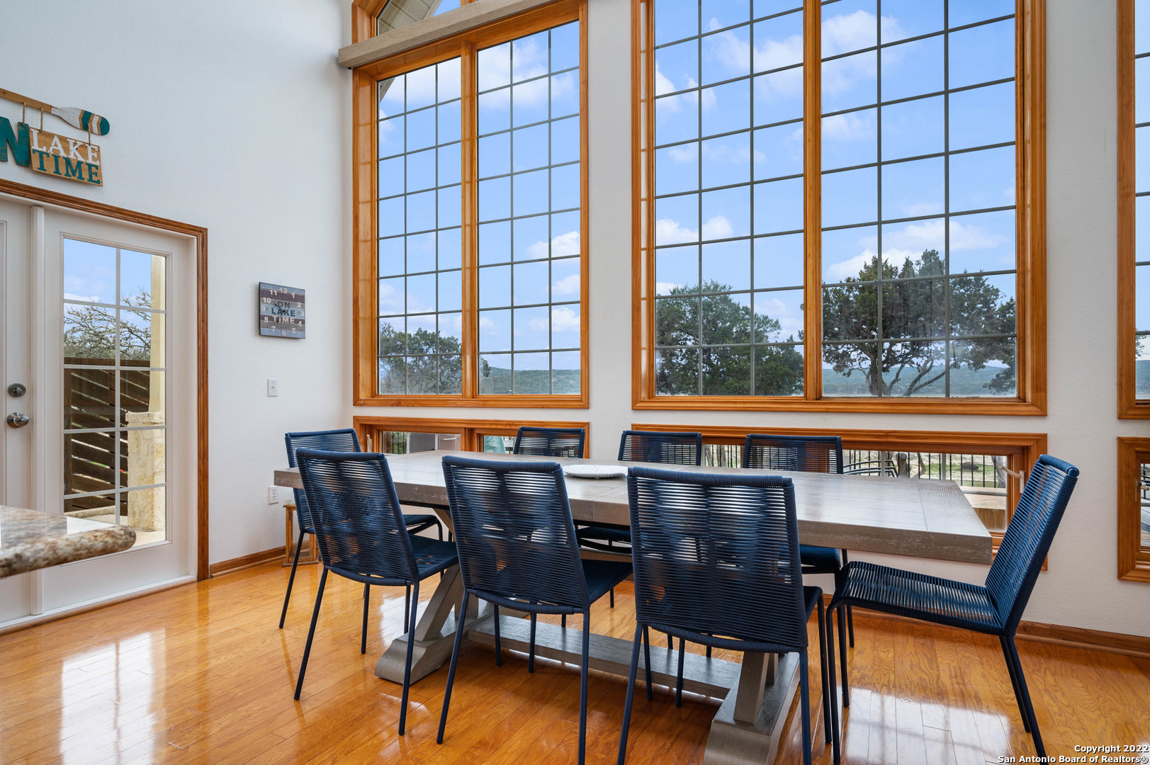 Dining area with a long table, dark woven chairs, and tall windows overlooking trees and distant hills.