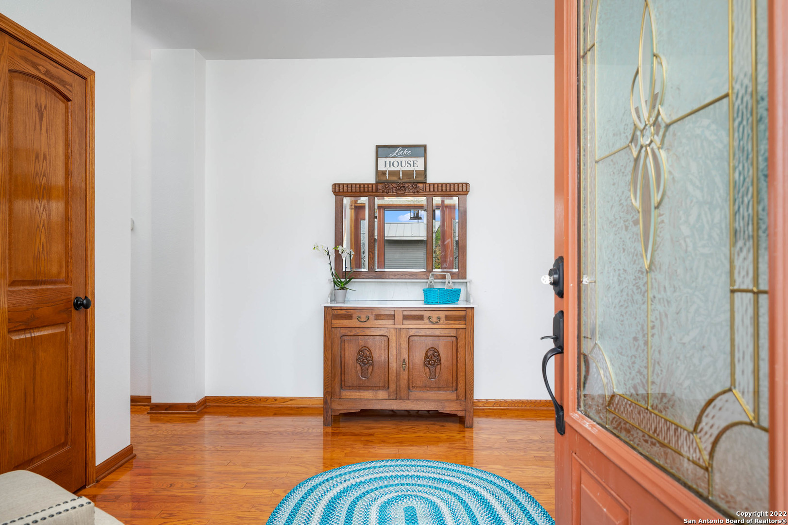 Entry foyer with polished wood floors, a carved console, blue rug, and open stained-glass door.