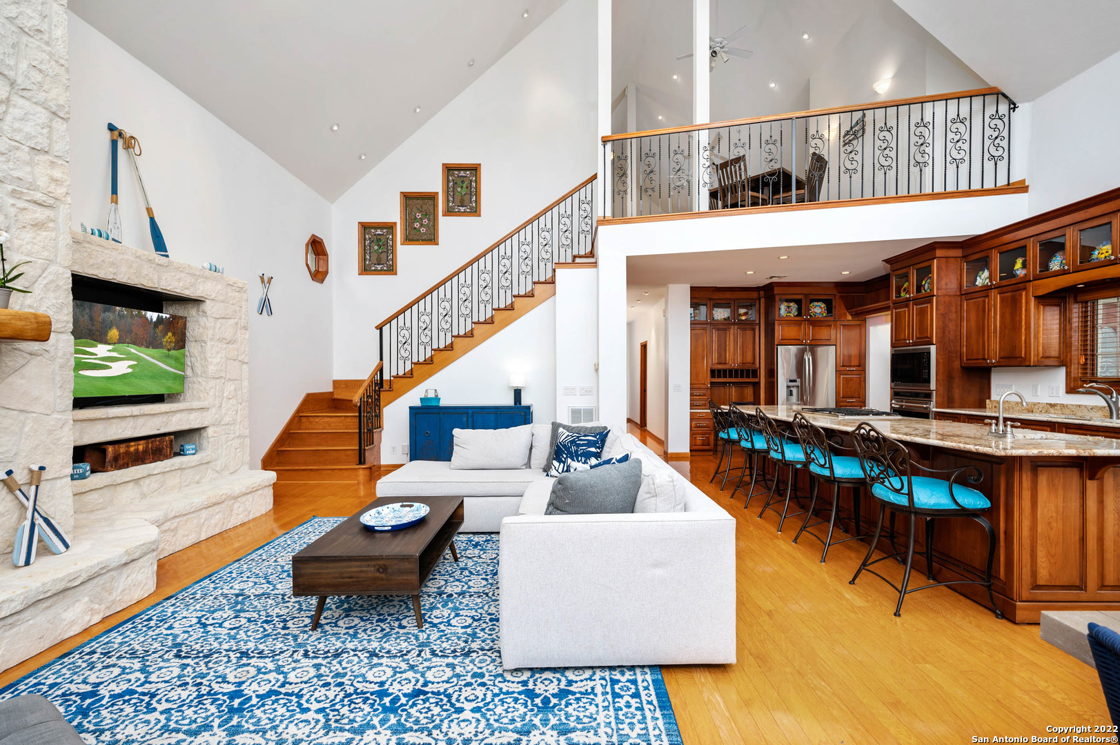 Furnished living room with a white stone fireplace, vaulted ceiling, staircase, and open kitchen.