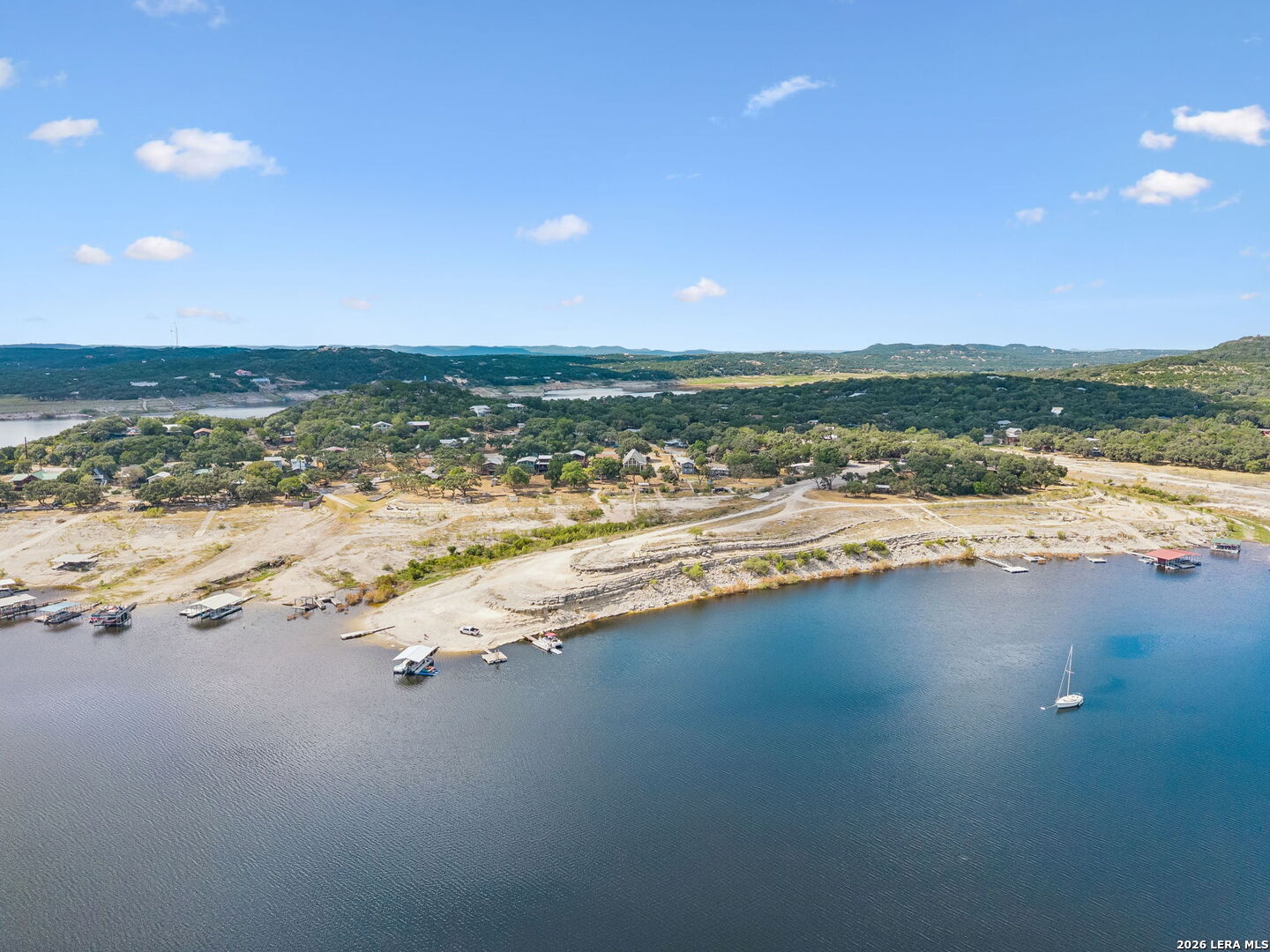 Aerial view of a lakeside neighborhood with shoreline docks, a sailboat, and wooded hills.