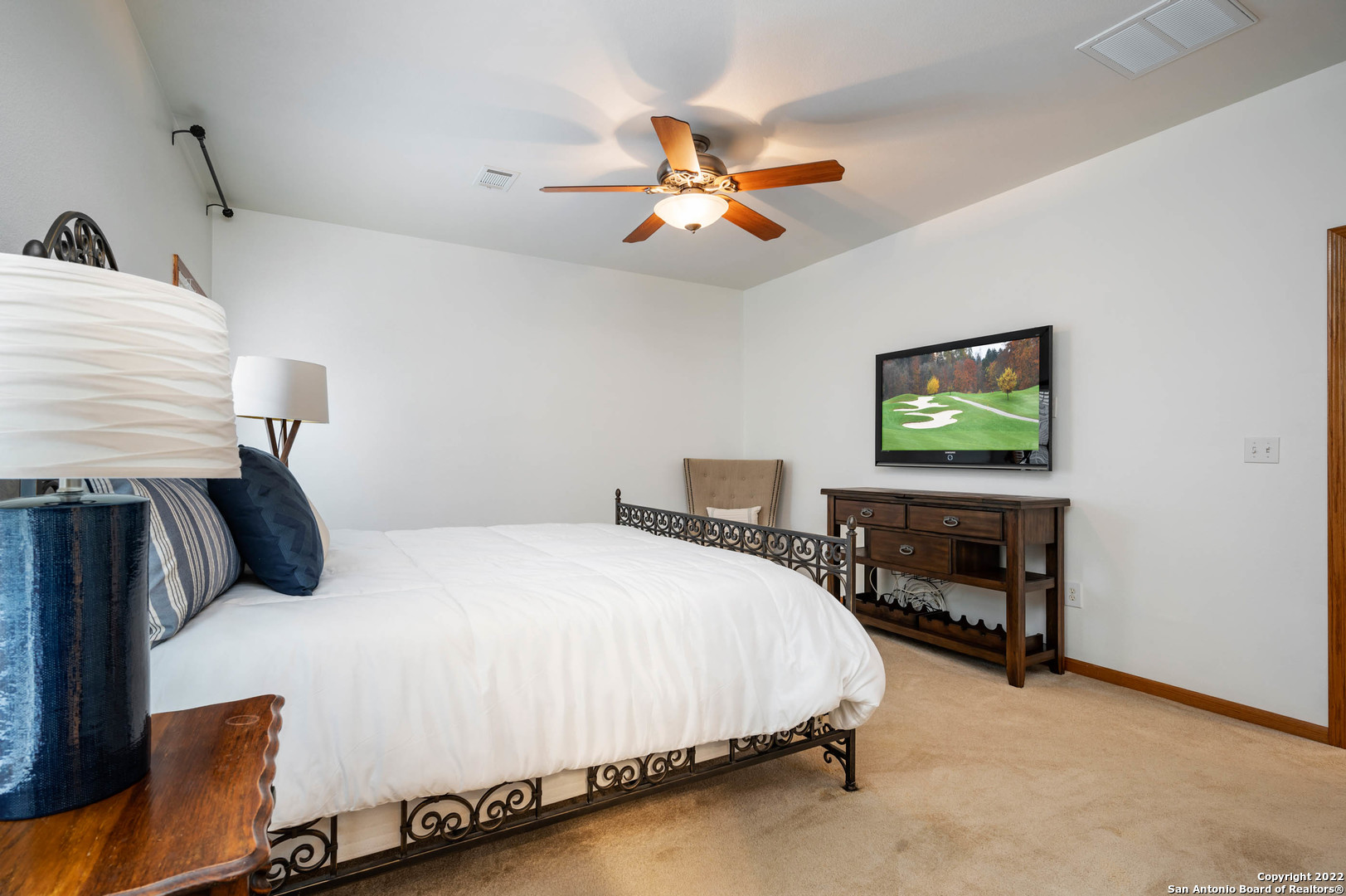 Bedroom with a white bed, ornate black metal frame, ceiling fan, and television showing a golf course.