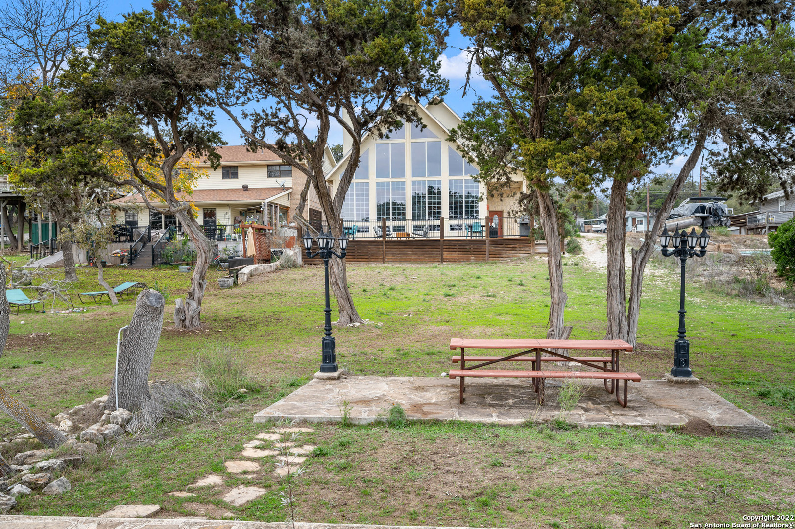 Home’s rear A-frame facade with tall windows beyond a shaded yard and stone patio with picnic table.