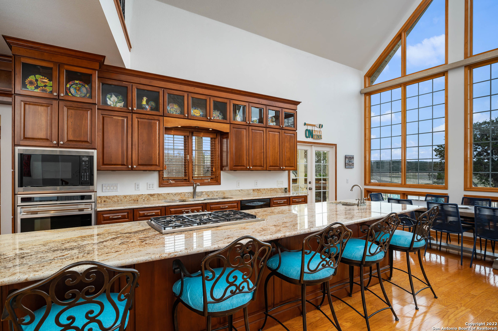 Kitchen with a granite island, turquoise bar stools, rich wood cabinetry, and tall windows.