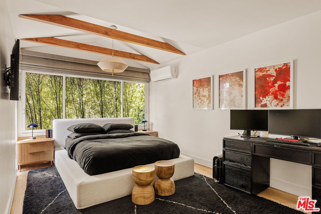 Staged bedroom with exposed wood beams, leafy views through a large window, and a dark desk with monitors.