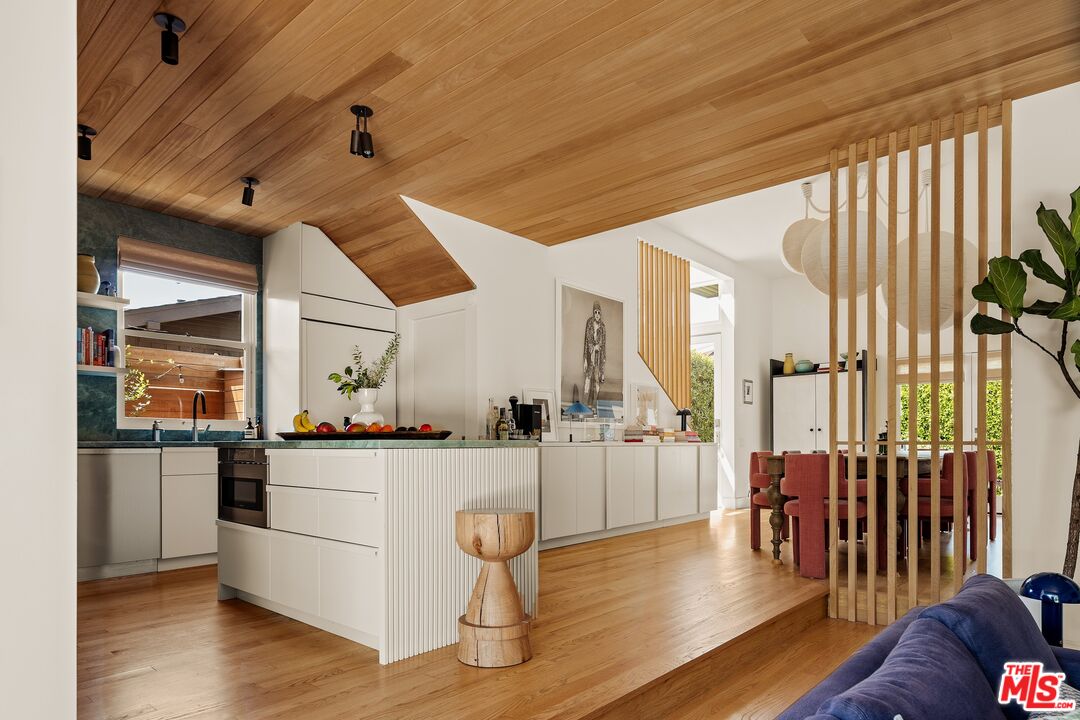 Open-plan kitchen with white cabinetry, fluted island, wood-paneled ceiling, and dining area behind a slatted divider.