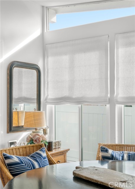 Dining area with woven chairs, blue patterned cushions, and tall windows with white shades.